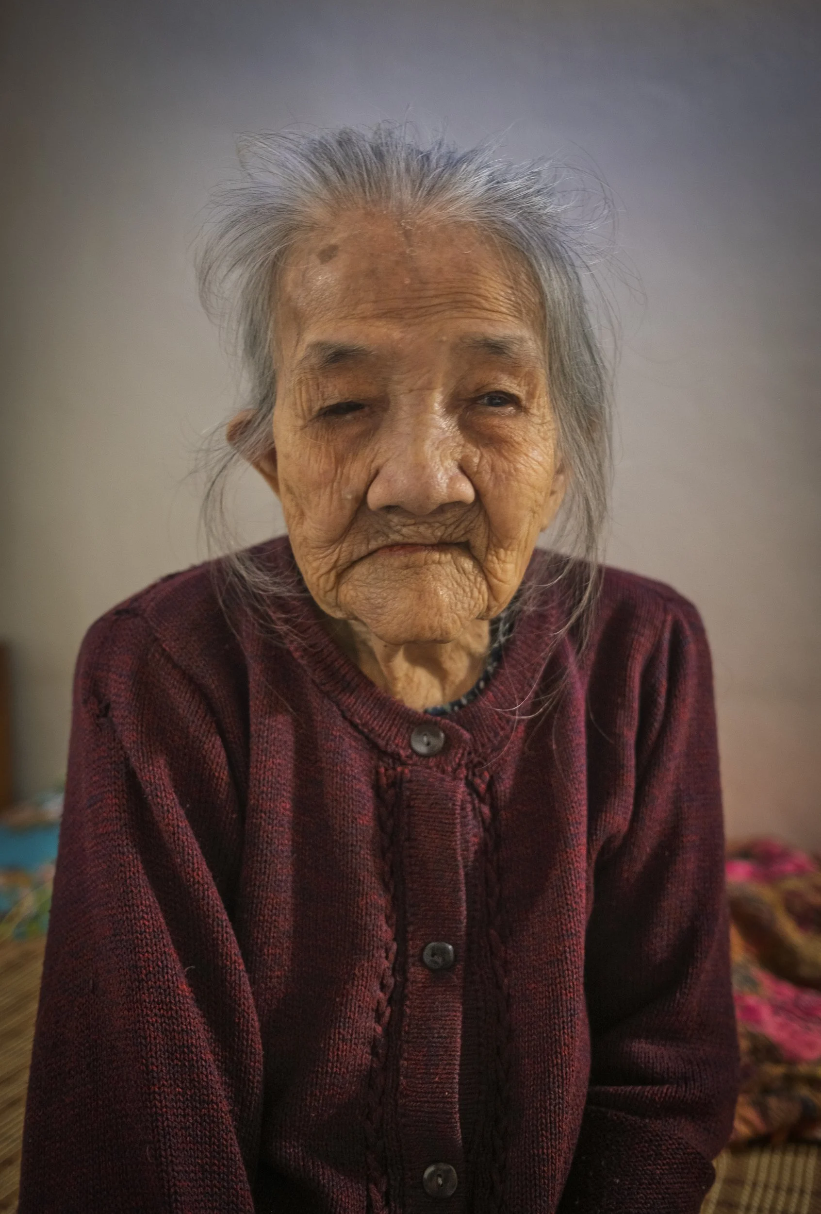 An elderly woman with gray hair, wearing a maroon sweater, sitting indoors.