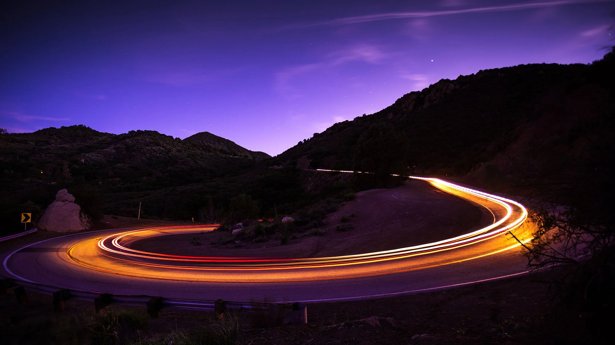 Long exposure photo of a winding mountain road at night with car light trails, purple sky with stars, and dark silhouetted hills.