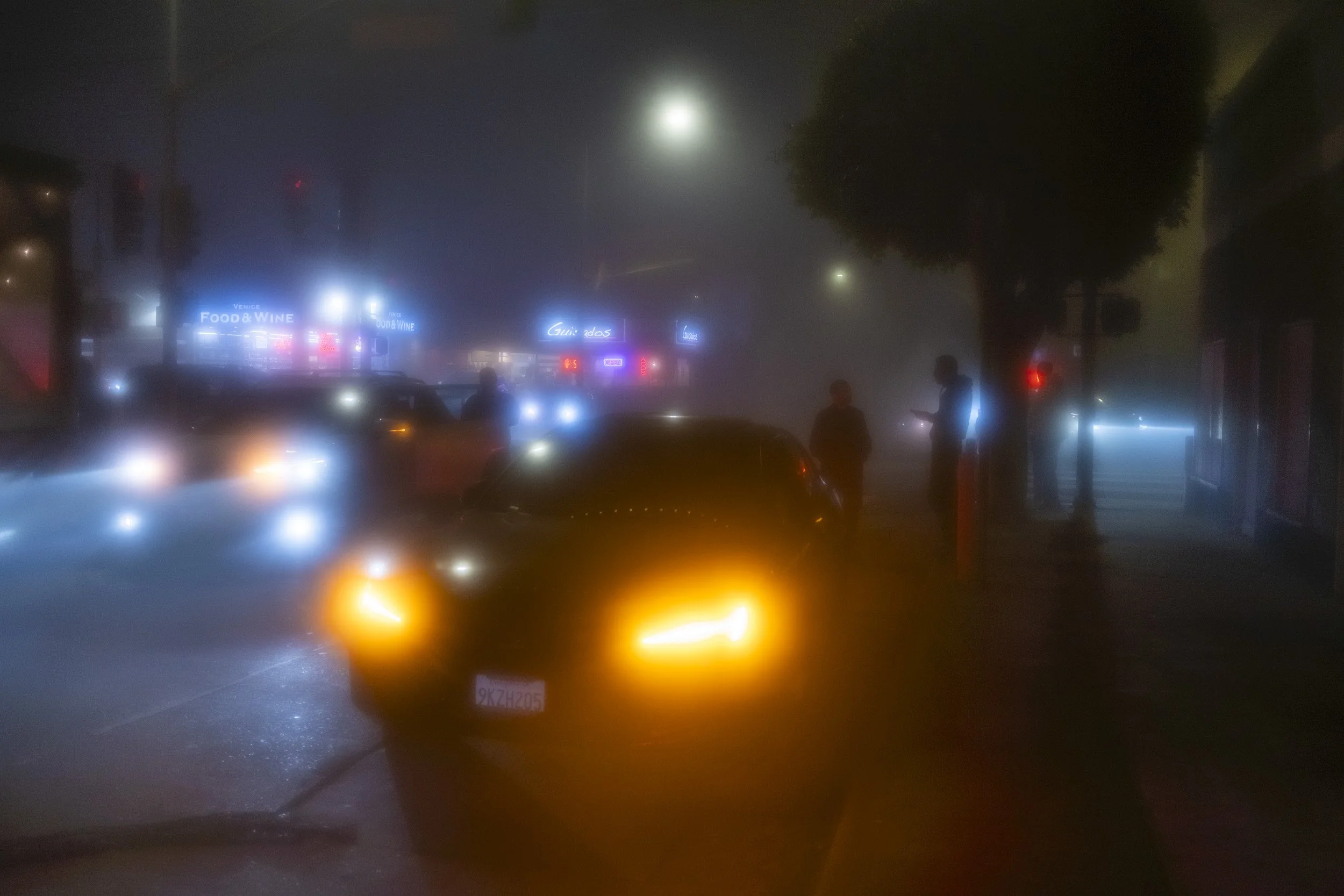 A foggy night scene on a city street with cars and pedestrians, illuminated by streetlights and neon signs, with limited visibility due to the thick fog.