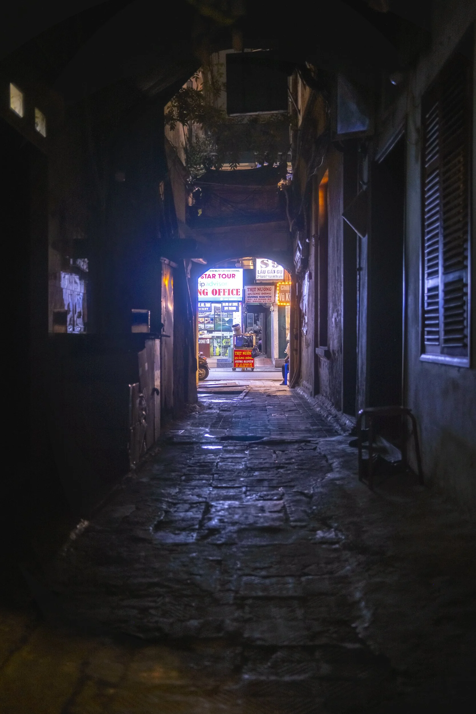 View of a narrow, dimly lit alleyway leading to a busy street with illuminated signs and storefronts in the distance.