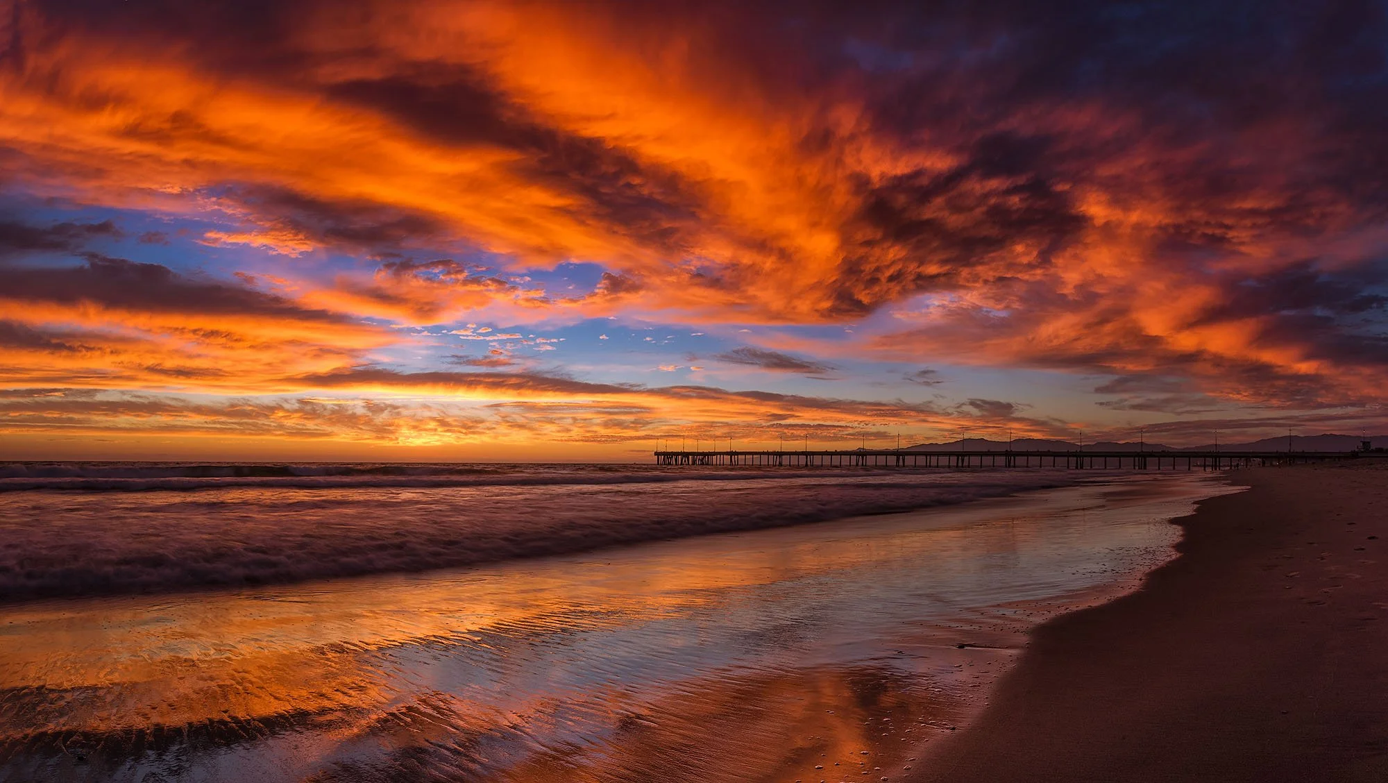 Colorful sunset over a beach with clouds in orange, pink, and purple hues reflecting on the wet sand and water, with a pier extending into the ocean in the distance.