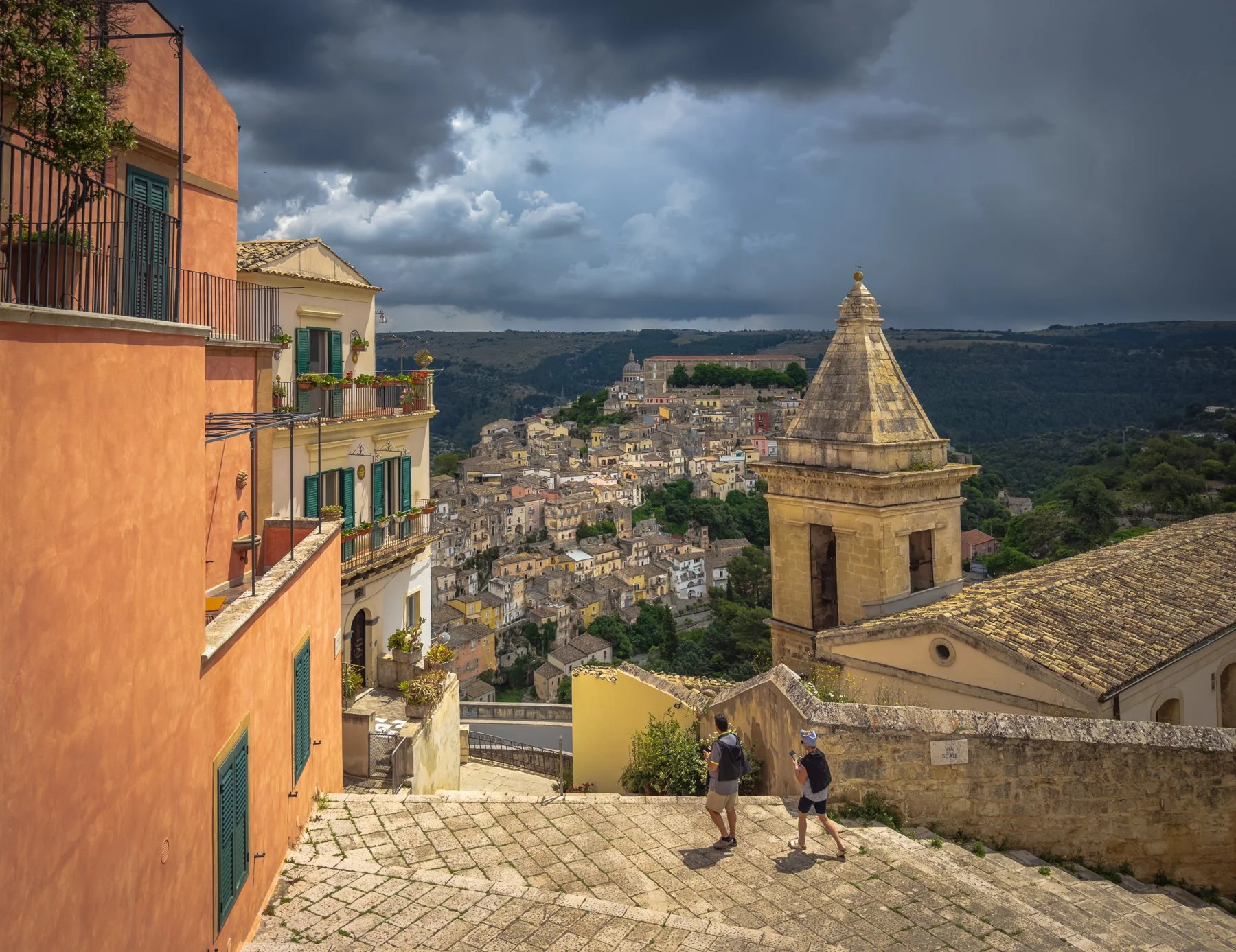 Overlooking a hillside town with colorful buildings, a church steeple, and two people walking on a cobblestone path under a cloudy sky.