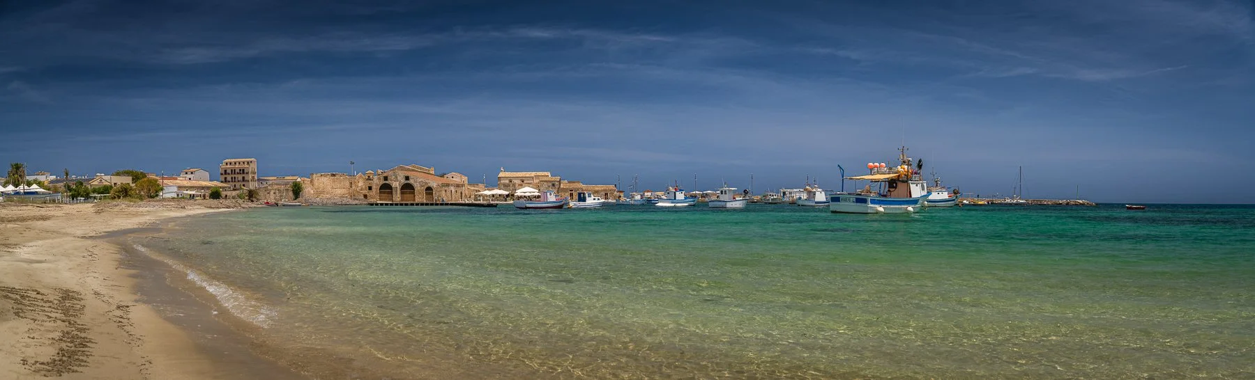 Beach with clear turquoise water, boats docked at a harbor, and historic buildings along the shoreline under a dark sky.