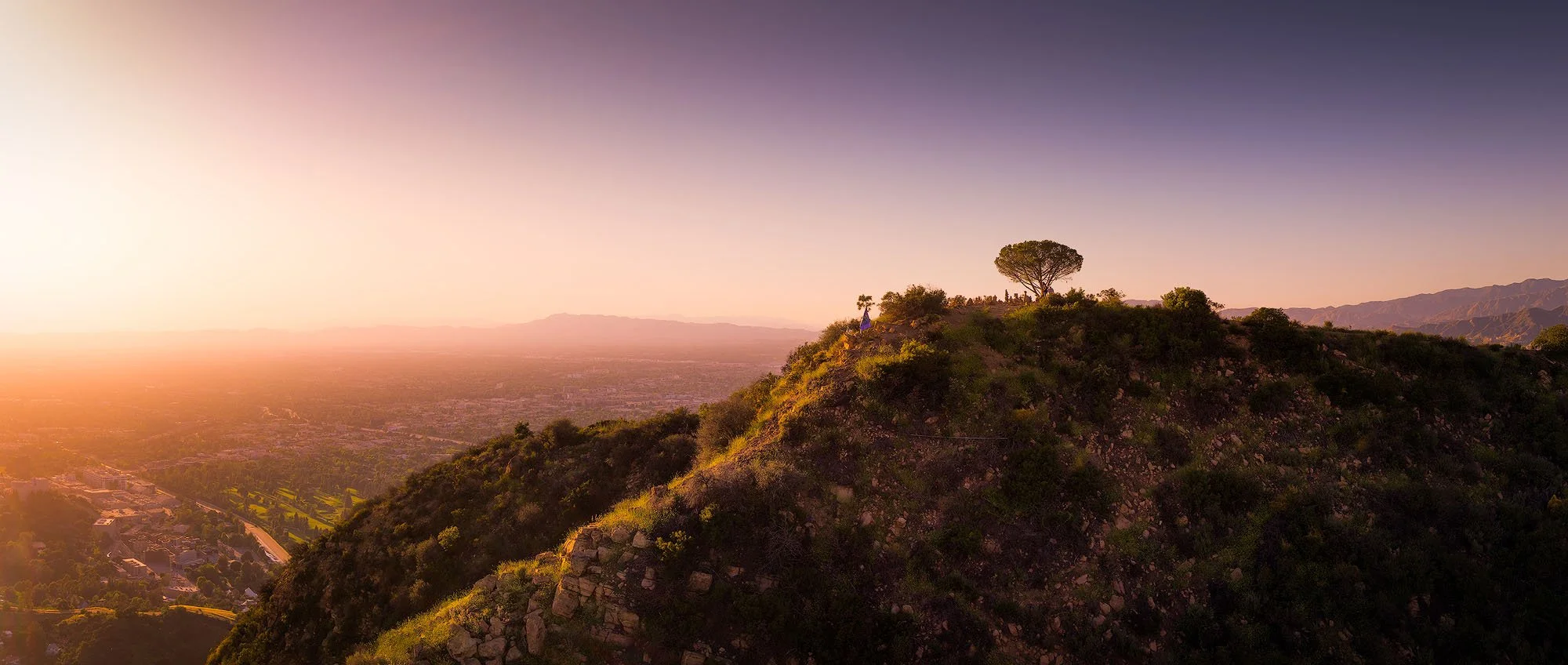 Sunset over a mountainous landscape with trees on a hilltop, distant city and mountain ranges in the background.