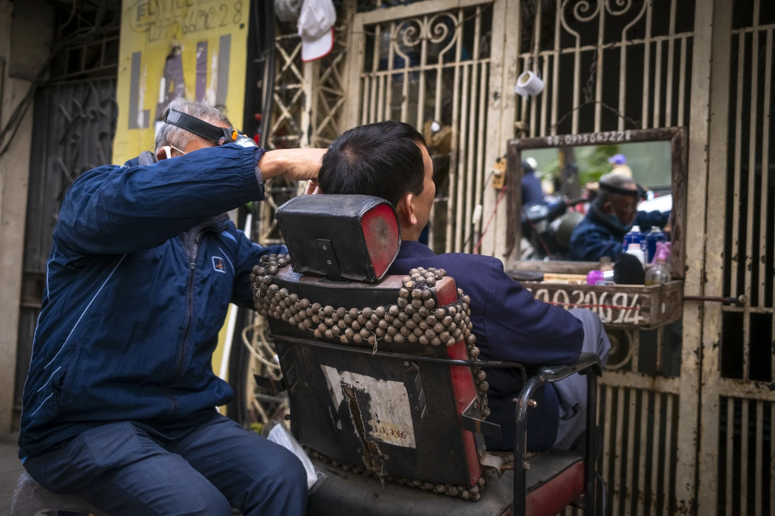 A man in a dark jacket is getting a haircut or grooming from a barber on the street seated in a wheelchair. The barber is wearing a blue jackets and is adjusting tools while looking at a mirror. The background shows a gated storefront with various it
