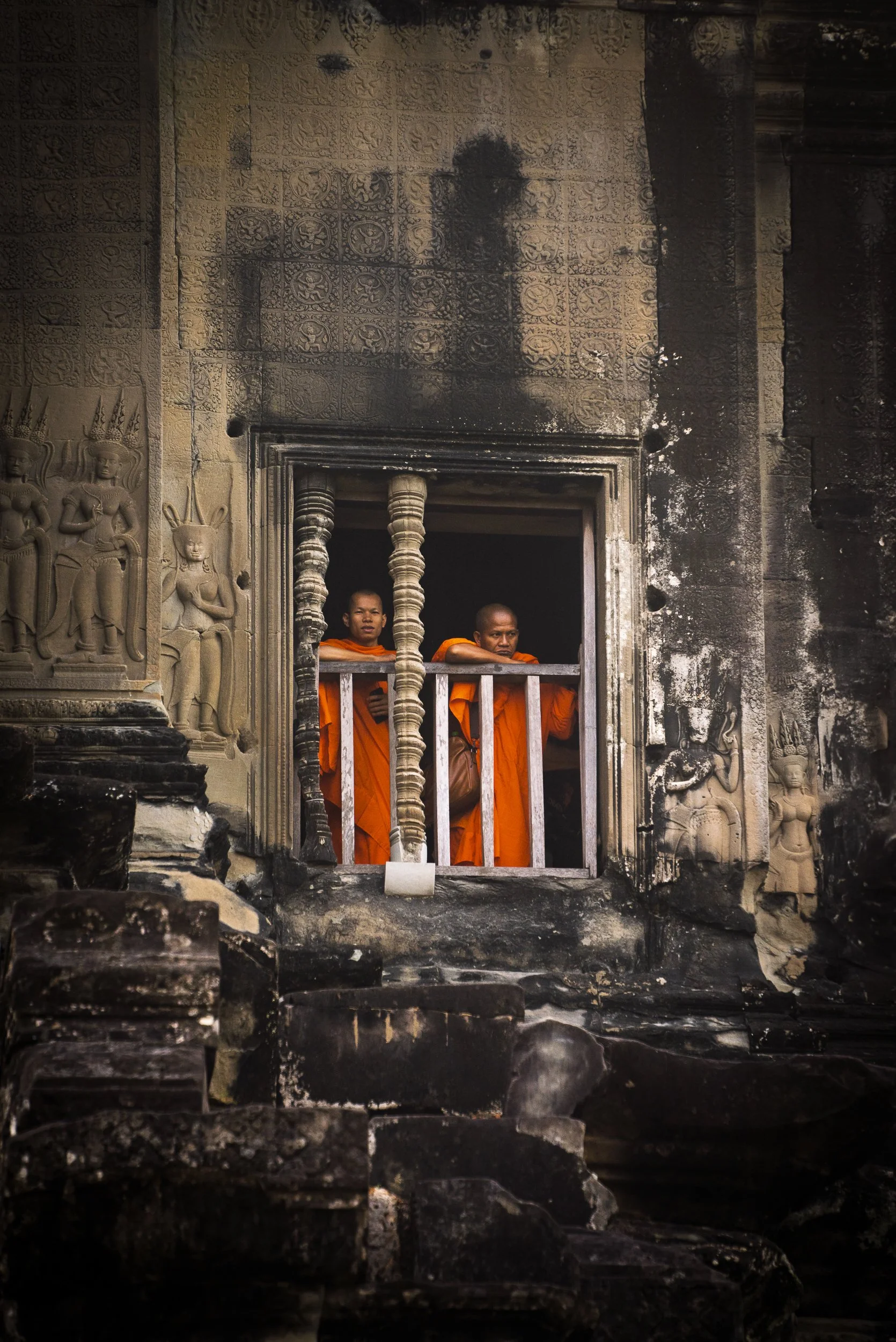 Two monks in orange robes standing behind a small barred window of an ancient, dark stone temple with intricate carvings and relief sculptures.