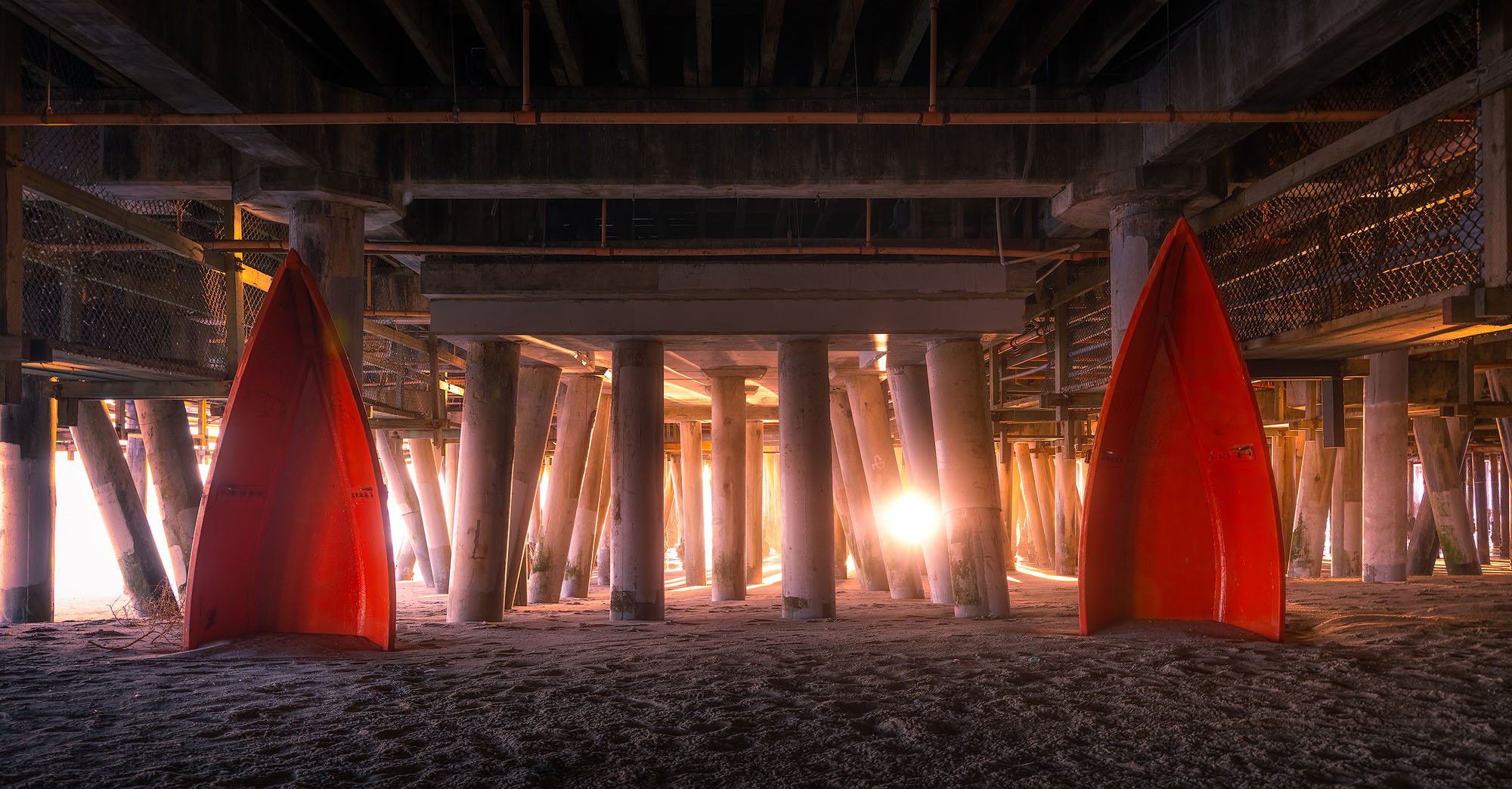 Underneath a pier at sunset with two red kayaks standing upright on the sandy ground.