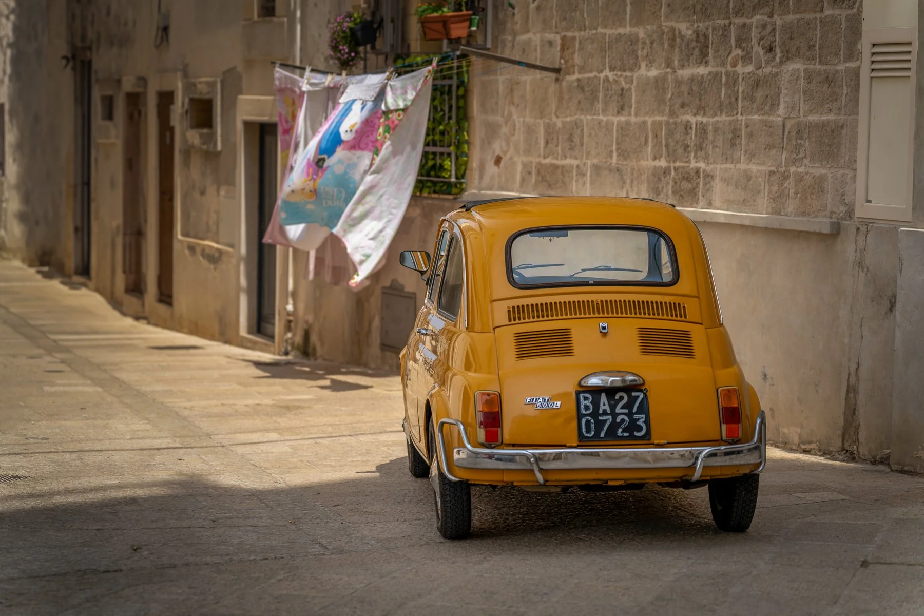 Yellow vintage Fiat 500 car parked on a narrow cobblestone street with clothes hanging on a line and a stone building wall in the background.