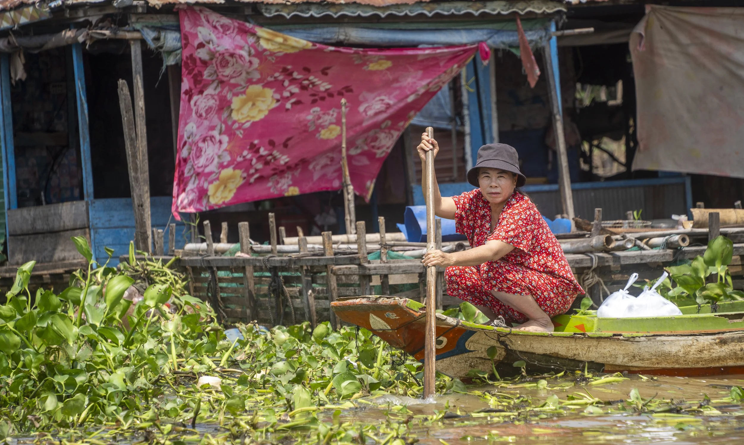 A woman in a red floral dress and gray hat paddles a small boat through a waterway filled with green aquatic plants. Behind her, there is a wooden structure with tarps and bamboo, suggesting a floating market or riverside dwelling in a rural area.