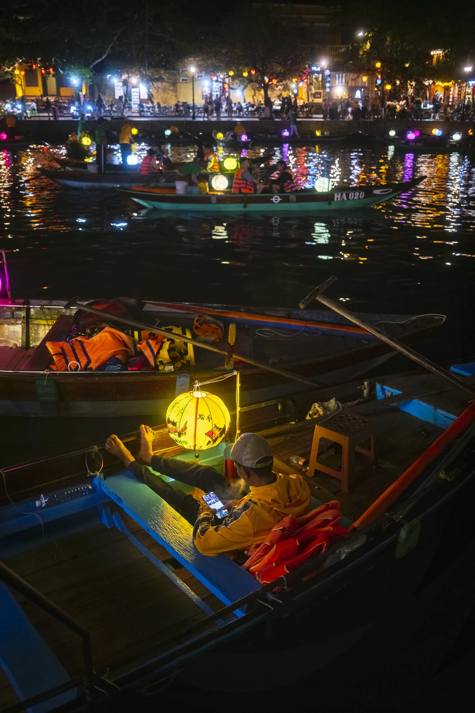 Person sitting in a boat at night, illuminated by a yellow lantern, looking at a smartphone with feet up on the boat's edge. Other boats with lanterns and a lively, lit-up riverside in the background.