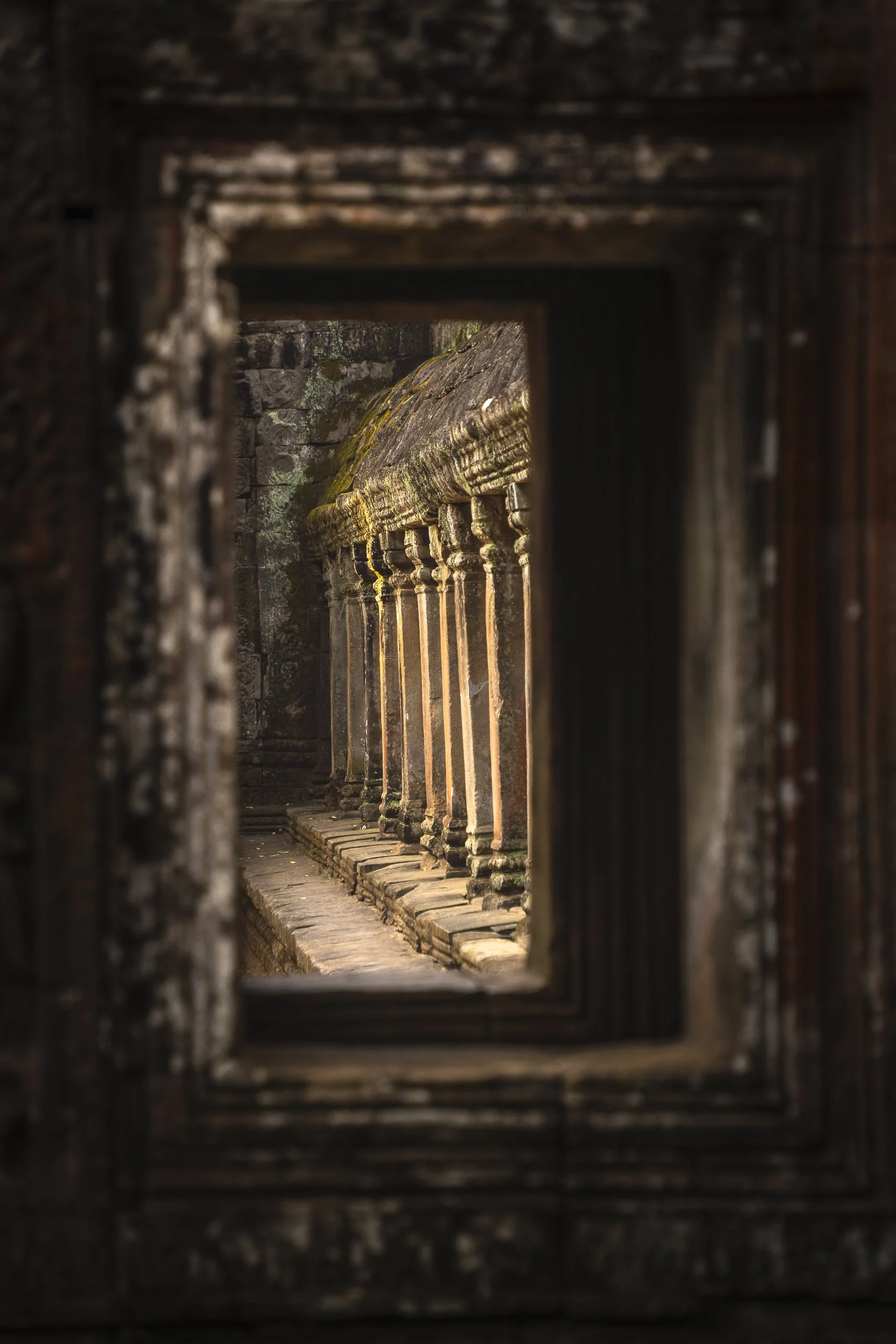 View through a small rectangular opening in an ancient stone wall, showcasing stone pillars with moss on top, in a historic temple or building.
