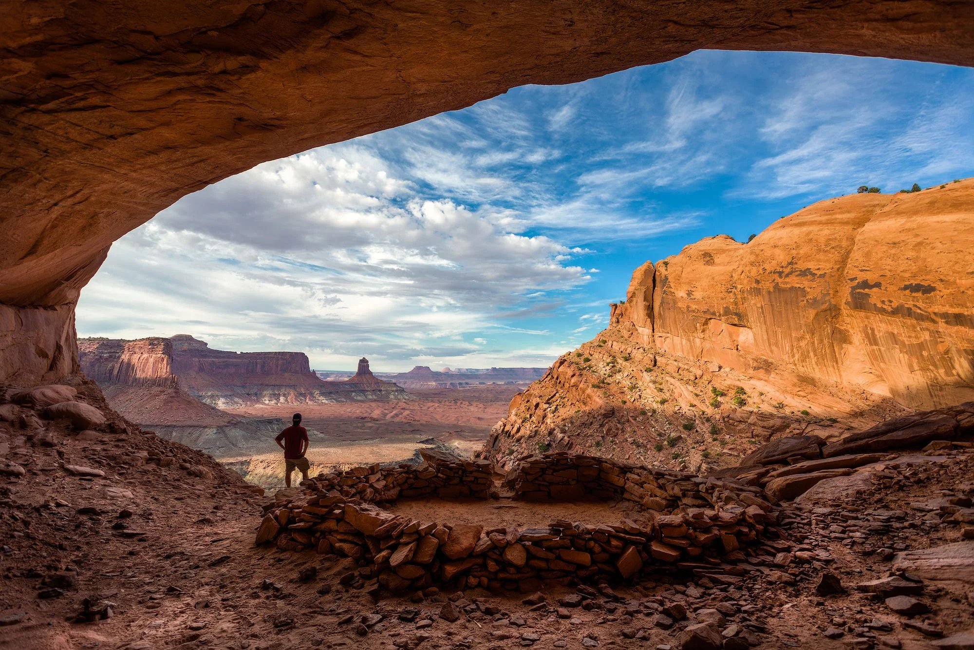 A person stands inside a rock alcove looking out over a desert landscape with mesas and rock formations under a partly cloudy sky.