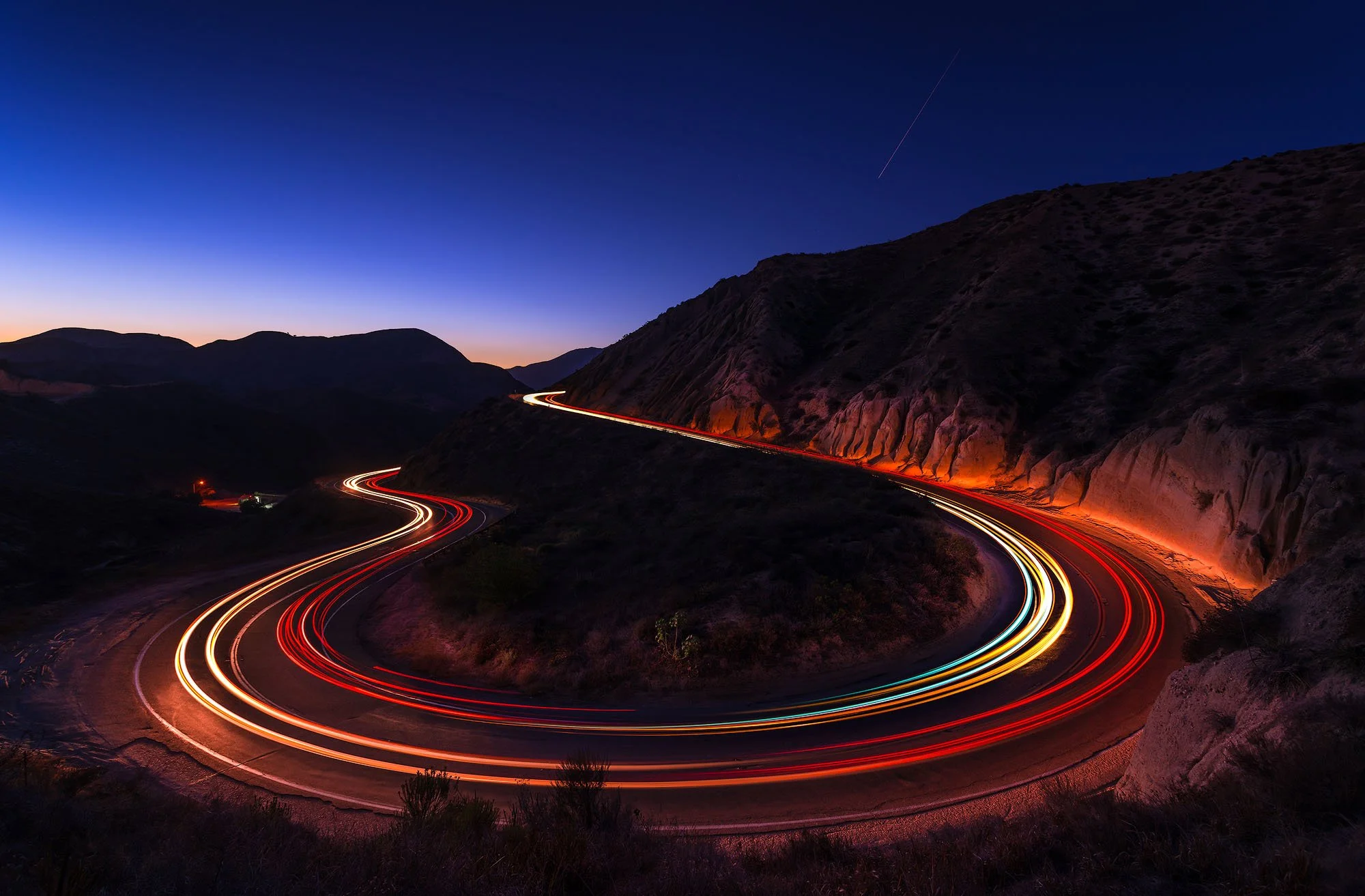 Winding mountain road at dusk with streaks of car lights and a dark blue sky.