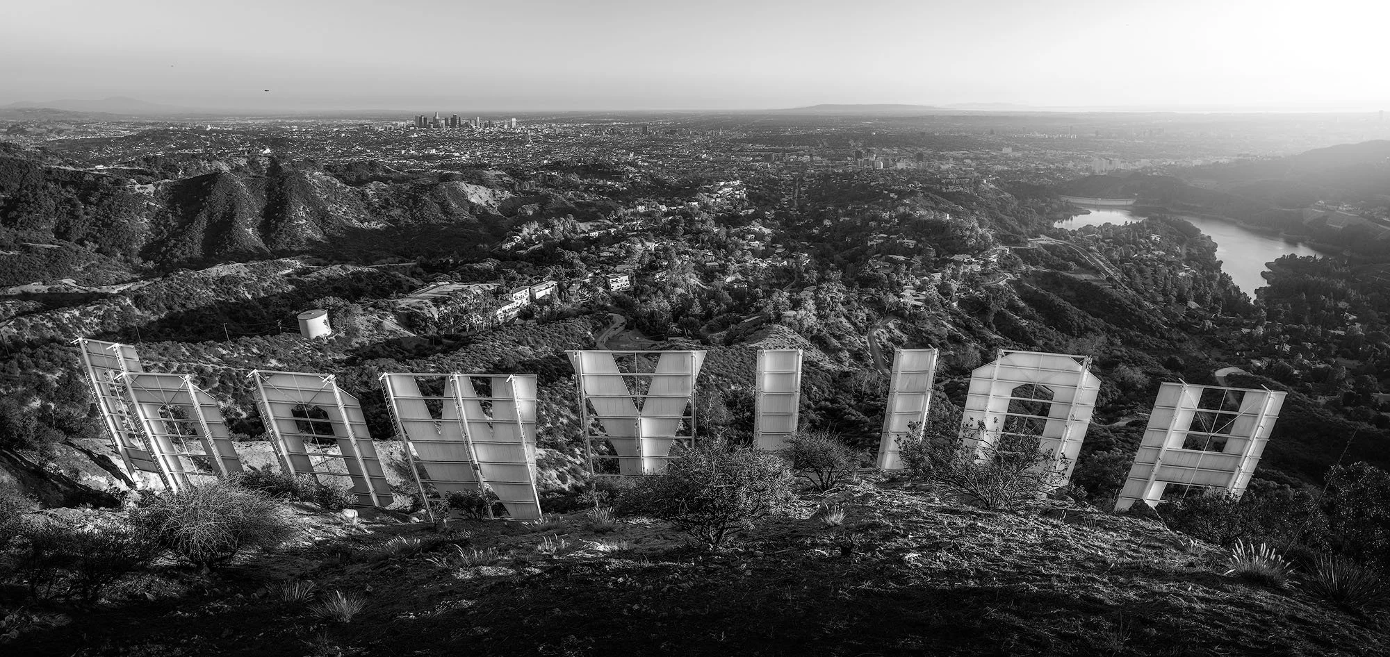 The Hollywood Sign in Los Angeles seen from a hillside with cityscape and river in the background, in black and white.