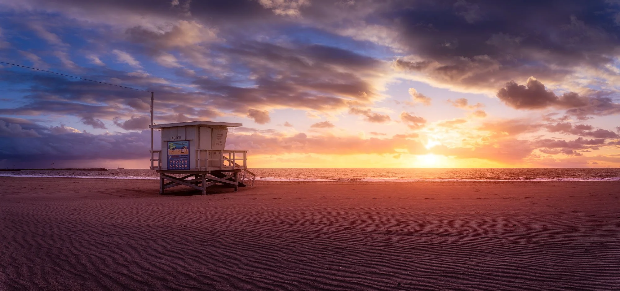 Sunset over a quiet beach with a lifeguard tower in the foreground and scattered clouds in the sky.