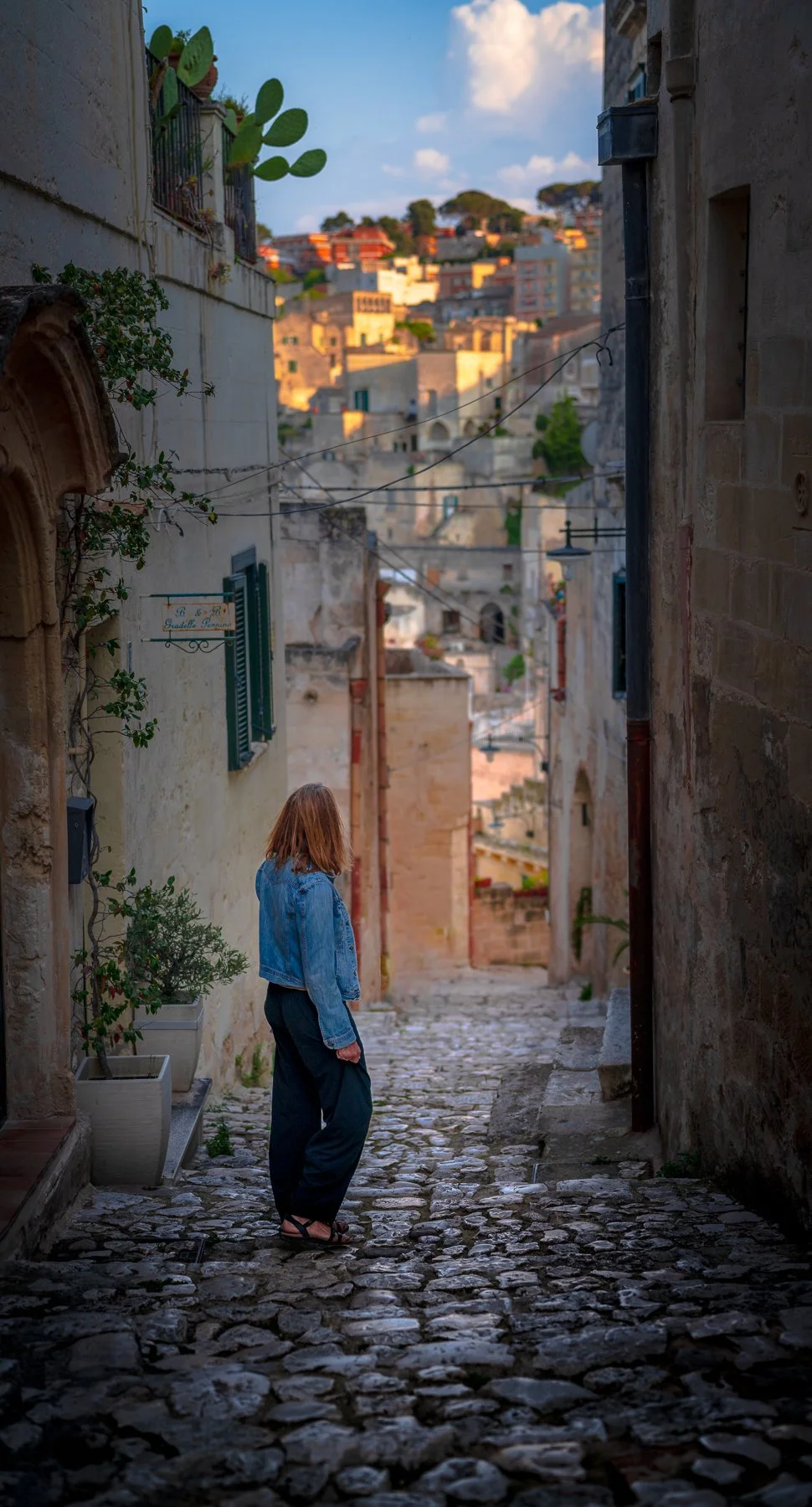 A woman with red hair wearing a blue denim jacket and black pants walks down a narrow cobblestone street in a historic European town with stone buildings and a hillside in the distance.