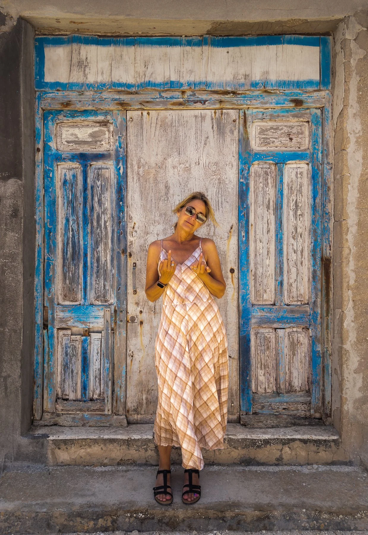 A woman wearing sunglasses and a flowy, checked maxi dress poses in front of a weathered, rustic wooden door with blue and white peeling paint. She is standing on a sidewalk, making a hand gesture with both hands.