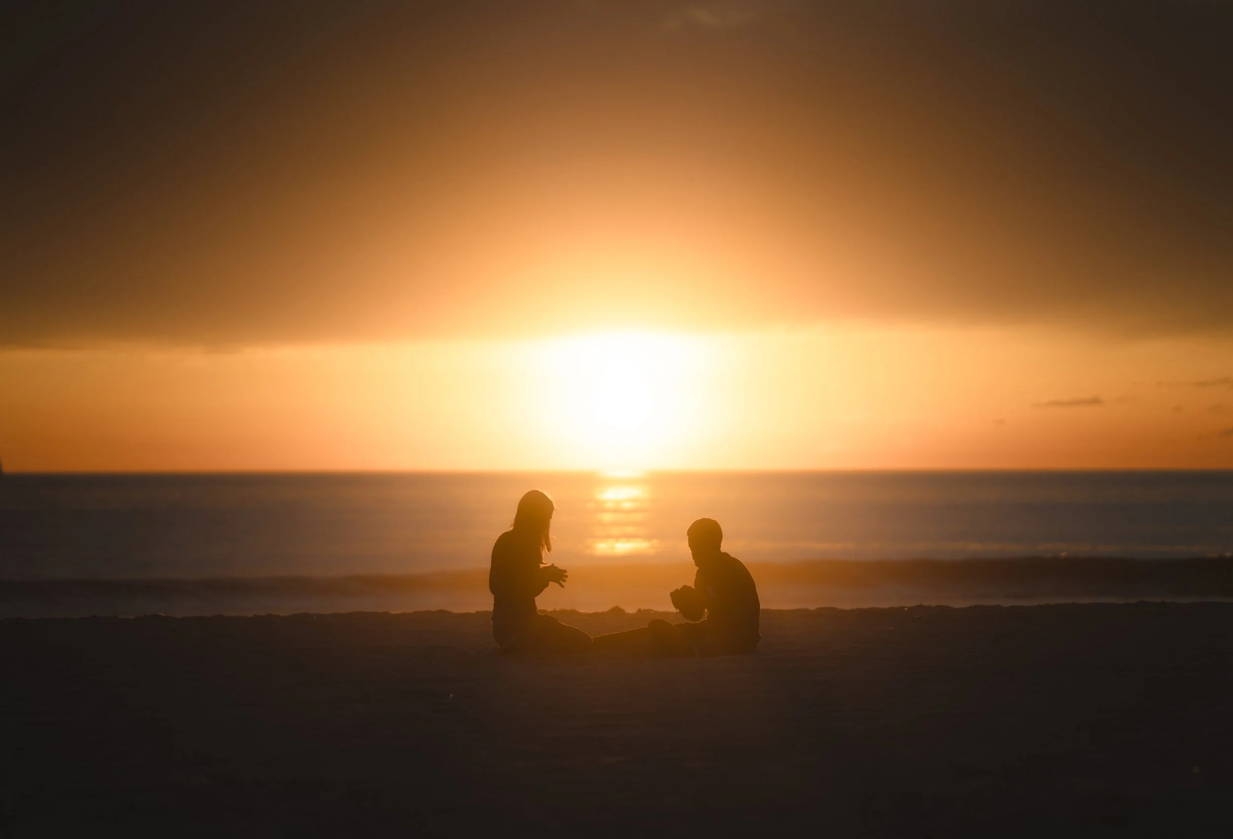 Silhouettes of two people sitting on the beach during a sunset, with the sun low on the horizon over the ocean.