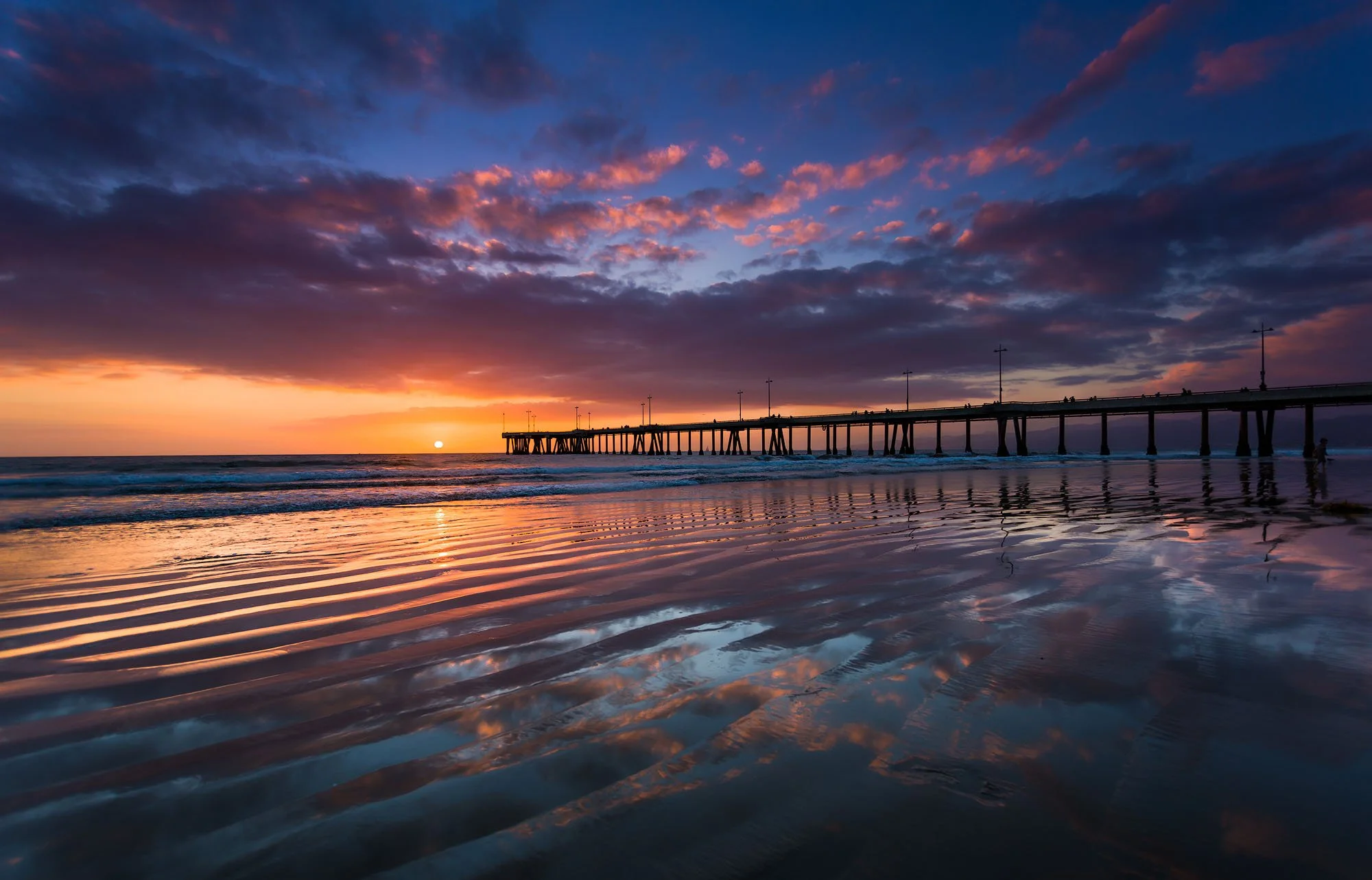 Sunset over the ocean with a pier extending into the water and reflected clouds on wet sand.