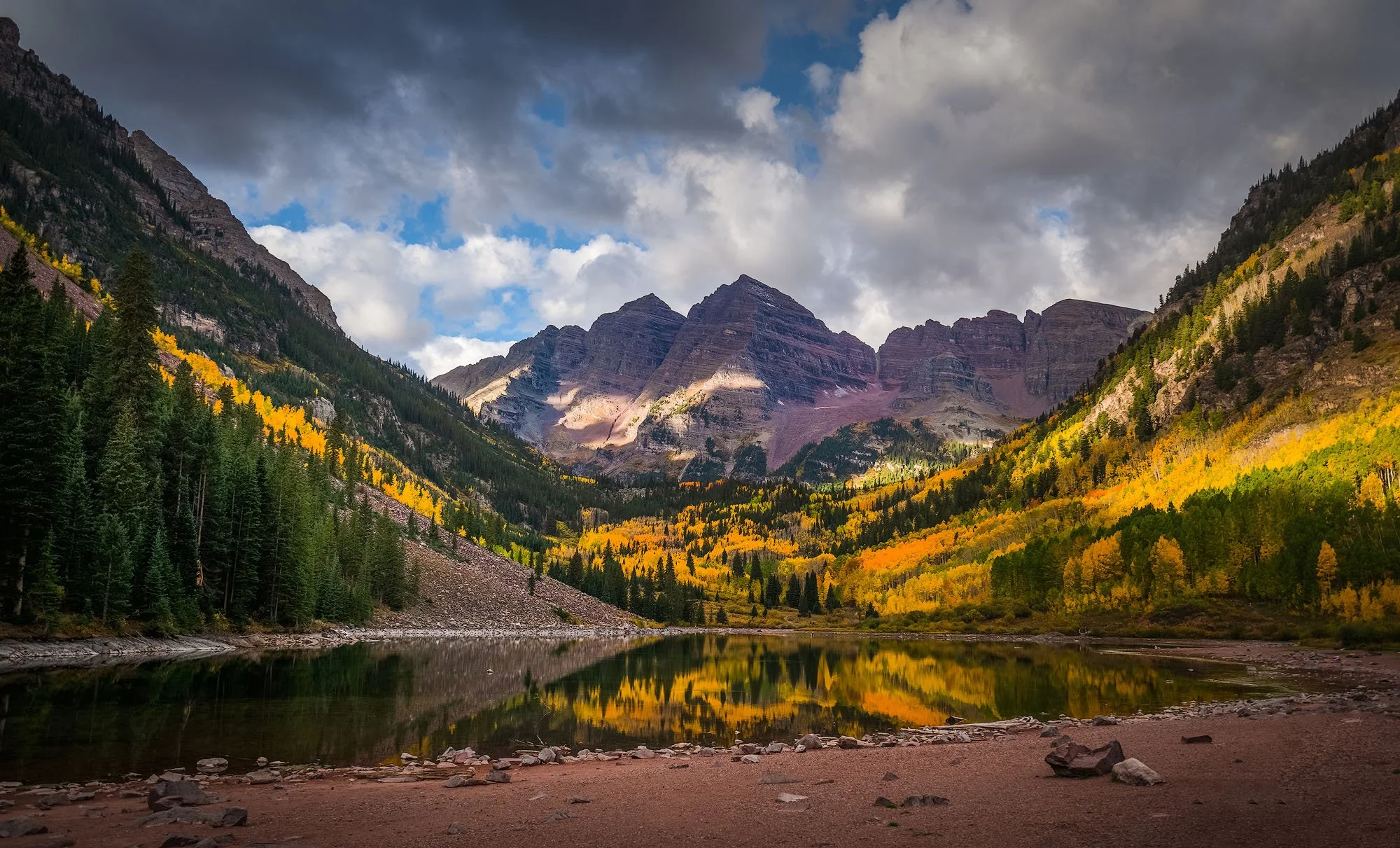 A mountain landscape with a lake in the foreground, surrounded by trees with fall colors and rugged mountain peaks under a partly cloudy sky.