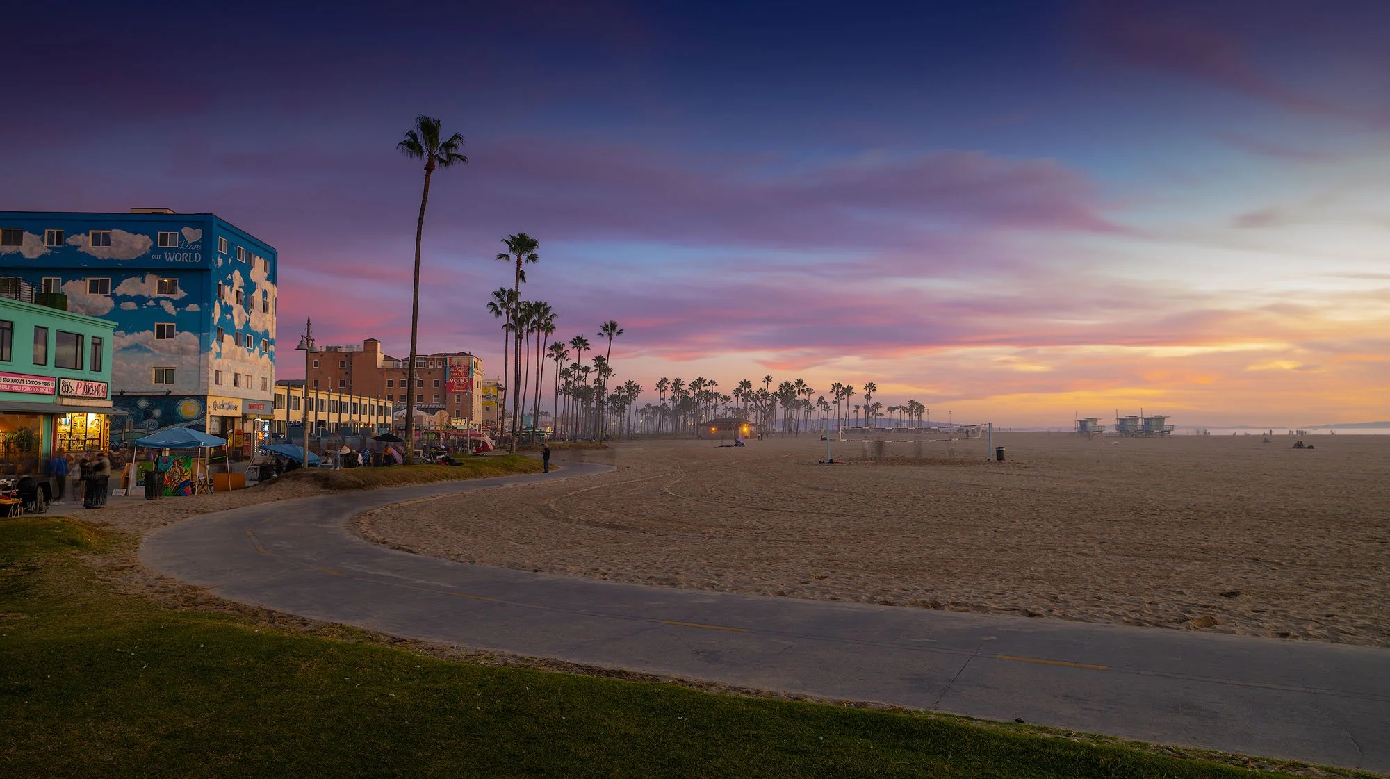 Sunset at the beach with colorful sky, palm trees, and buildings along the shoreline.