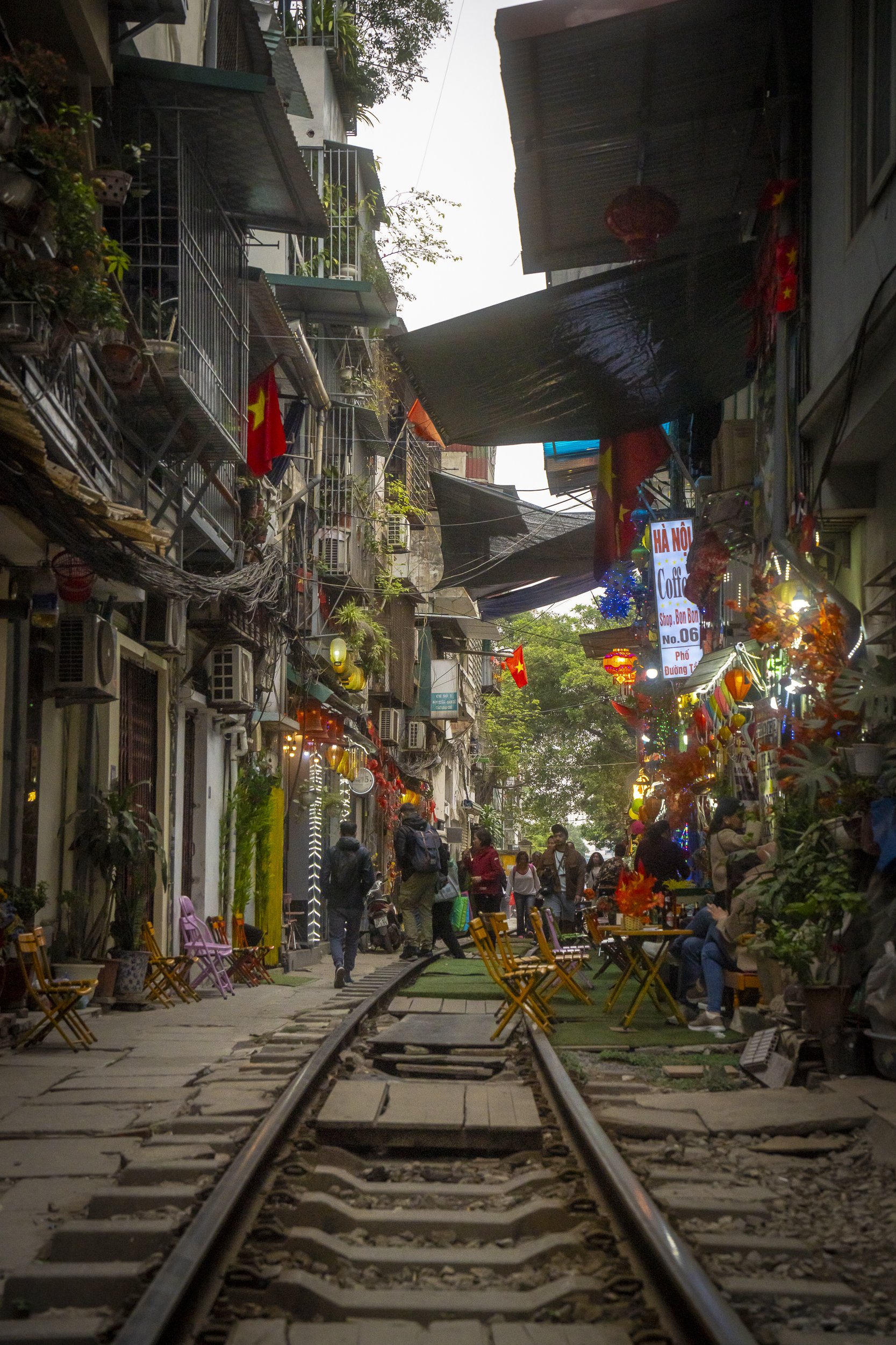 A bustling street scene in an urban neighborhood with shops, cafes, and outdoor seating alongside a railway track, decorated with flags and colorful lights, with people walking and sitting.