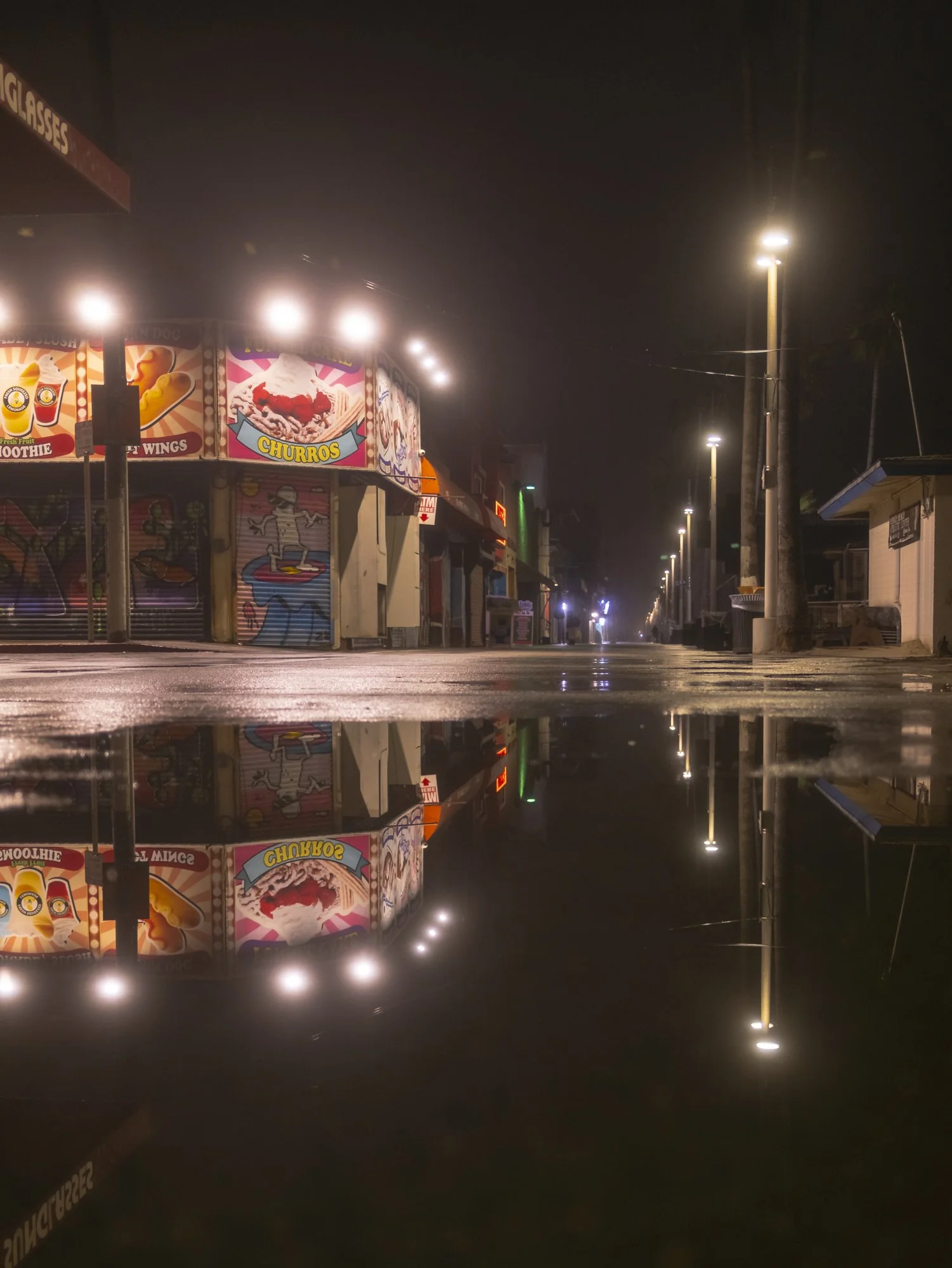 Nighttime street scene with closed shops and colorful illuminated signs, reflected in a large puddle on the wet pavement.