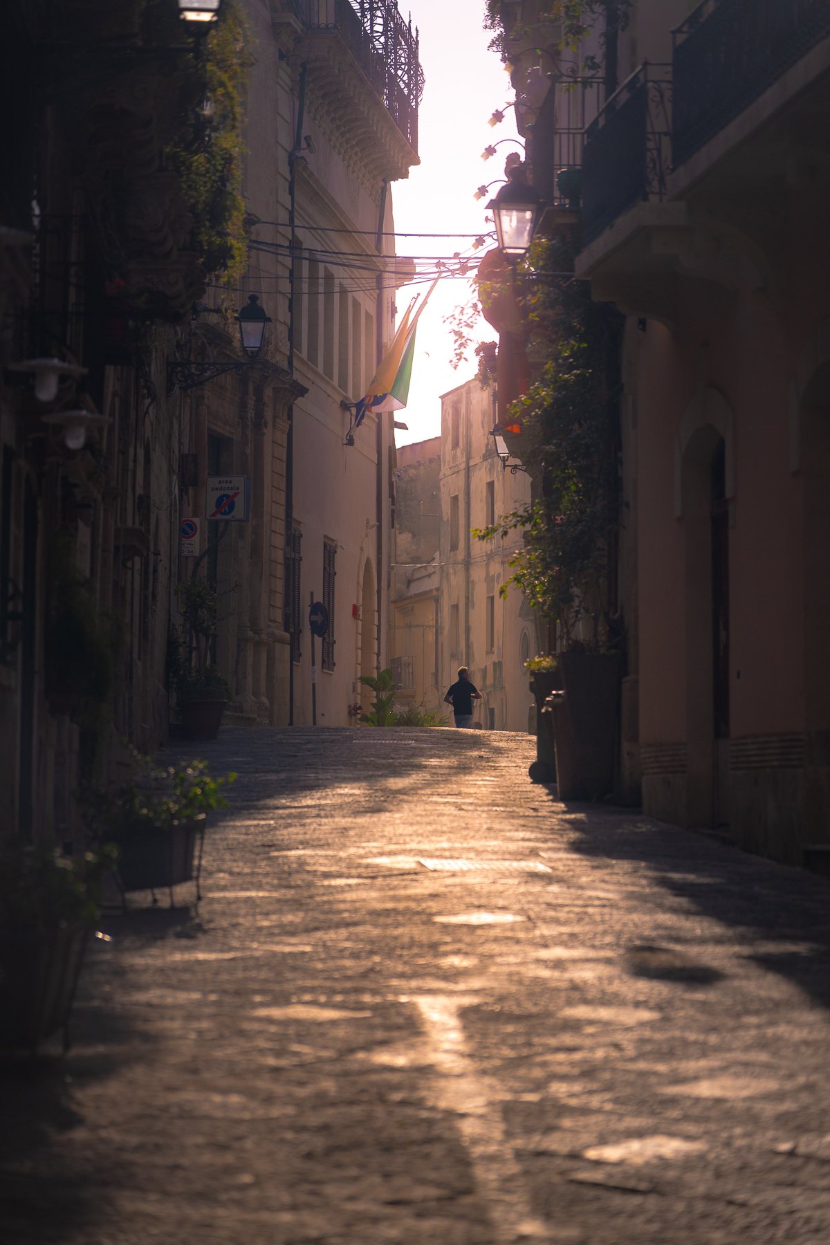 A narrow, cobblestone street in a European city during sunset, with a person walking away from the camera and buildings with balconies and potted plants on either side.