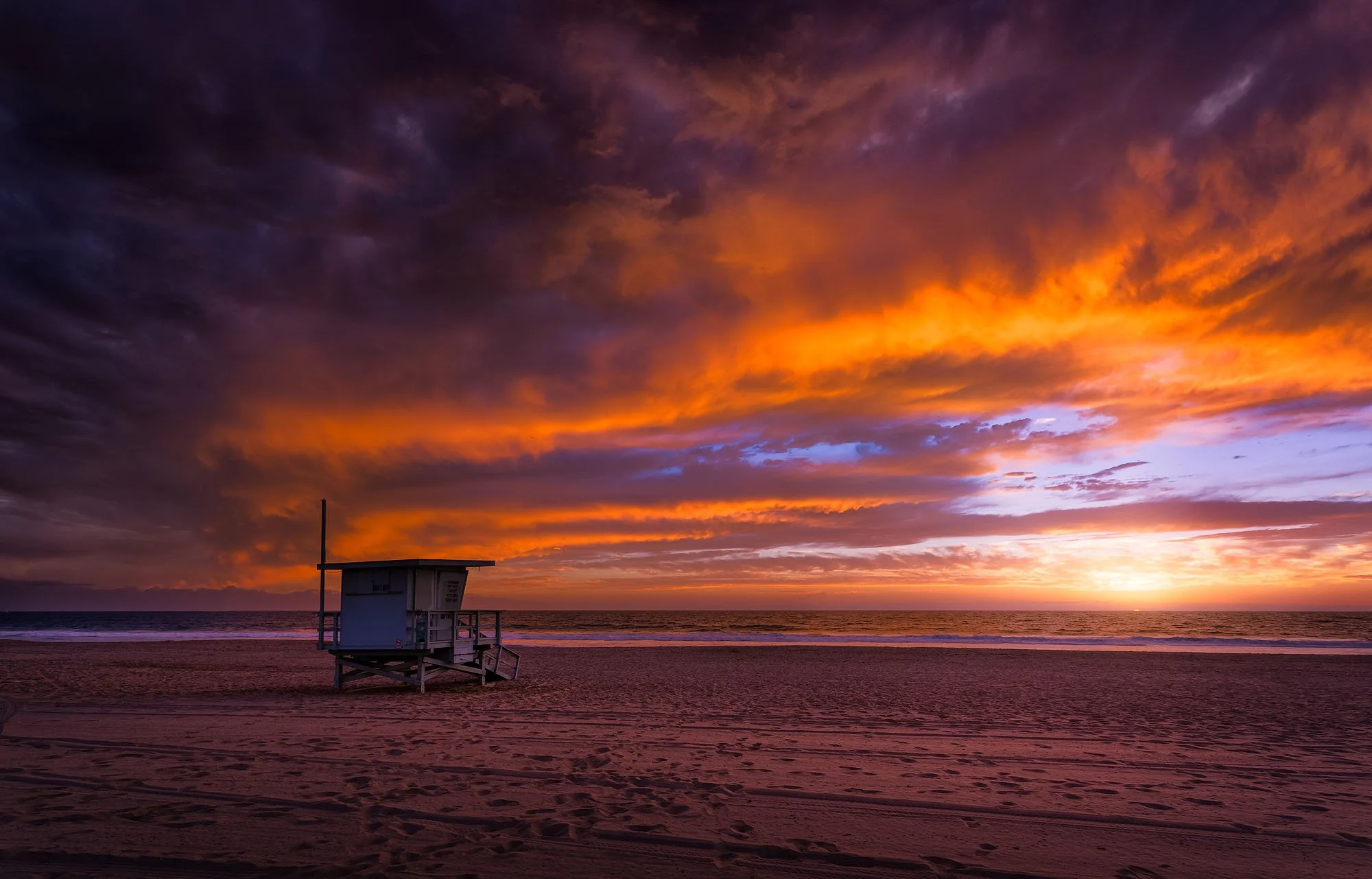 A lifeguard tower on the beach at sunset with a vibrant orange, purple, and pink sky filled with clouds over the ocean.