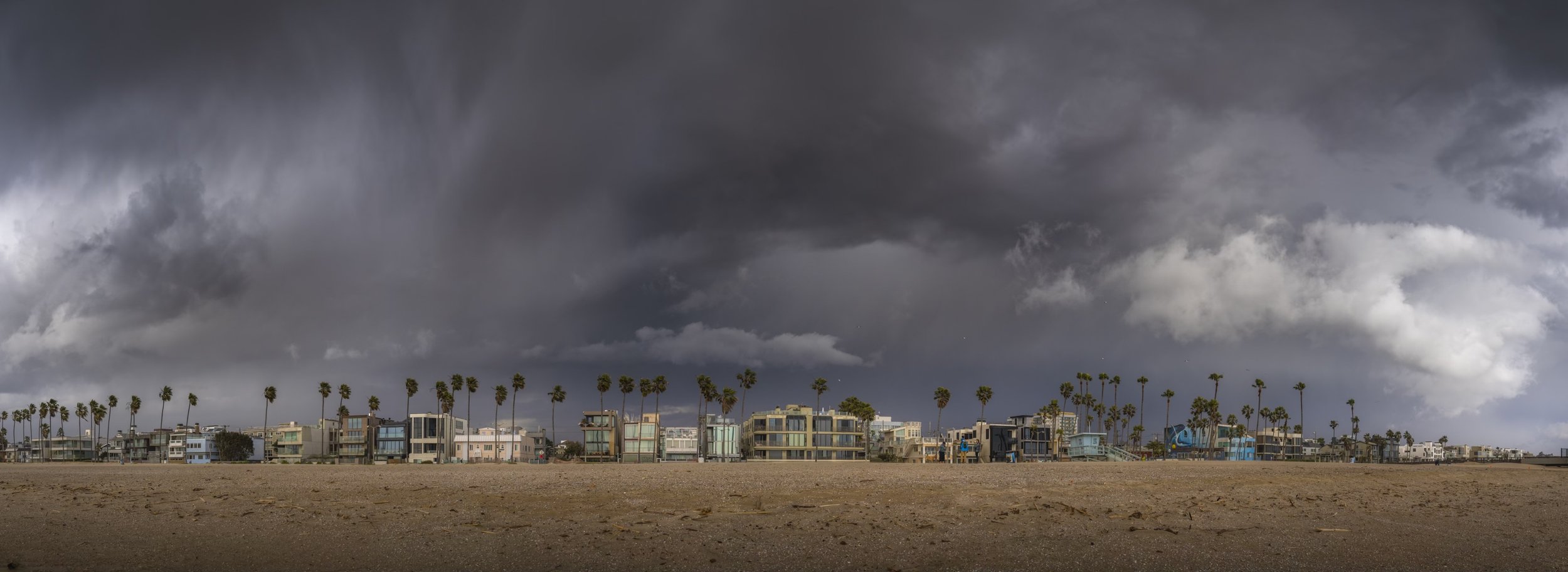 Overcast sky with dark storm clouds over a row of beach houses and palm trees along the shoreline.