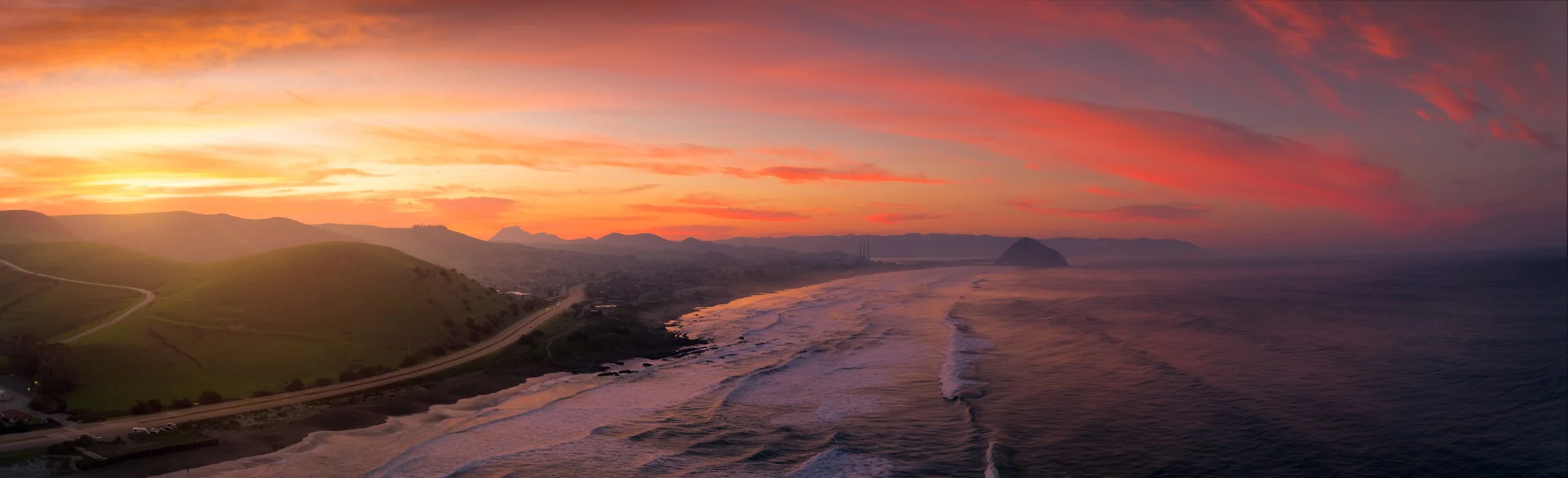 Sunset over a coastal landscape with hills, a winding road, and the ocean with waves