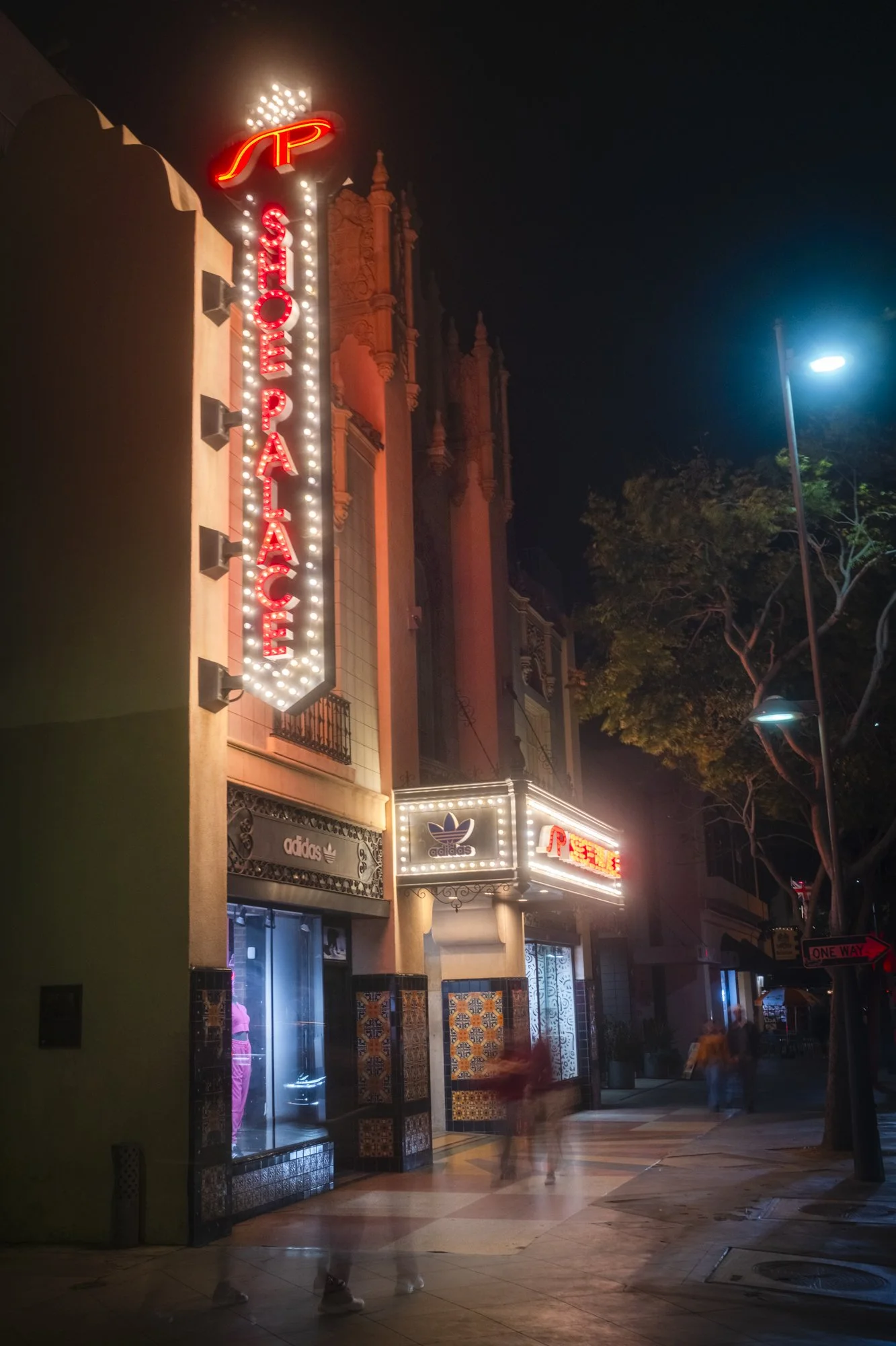 Night view of a theater with illuminated neon signs that say "The Palace" and the Adidas logo, with blurred pedestrians walking by on the sidewalk.