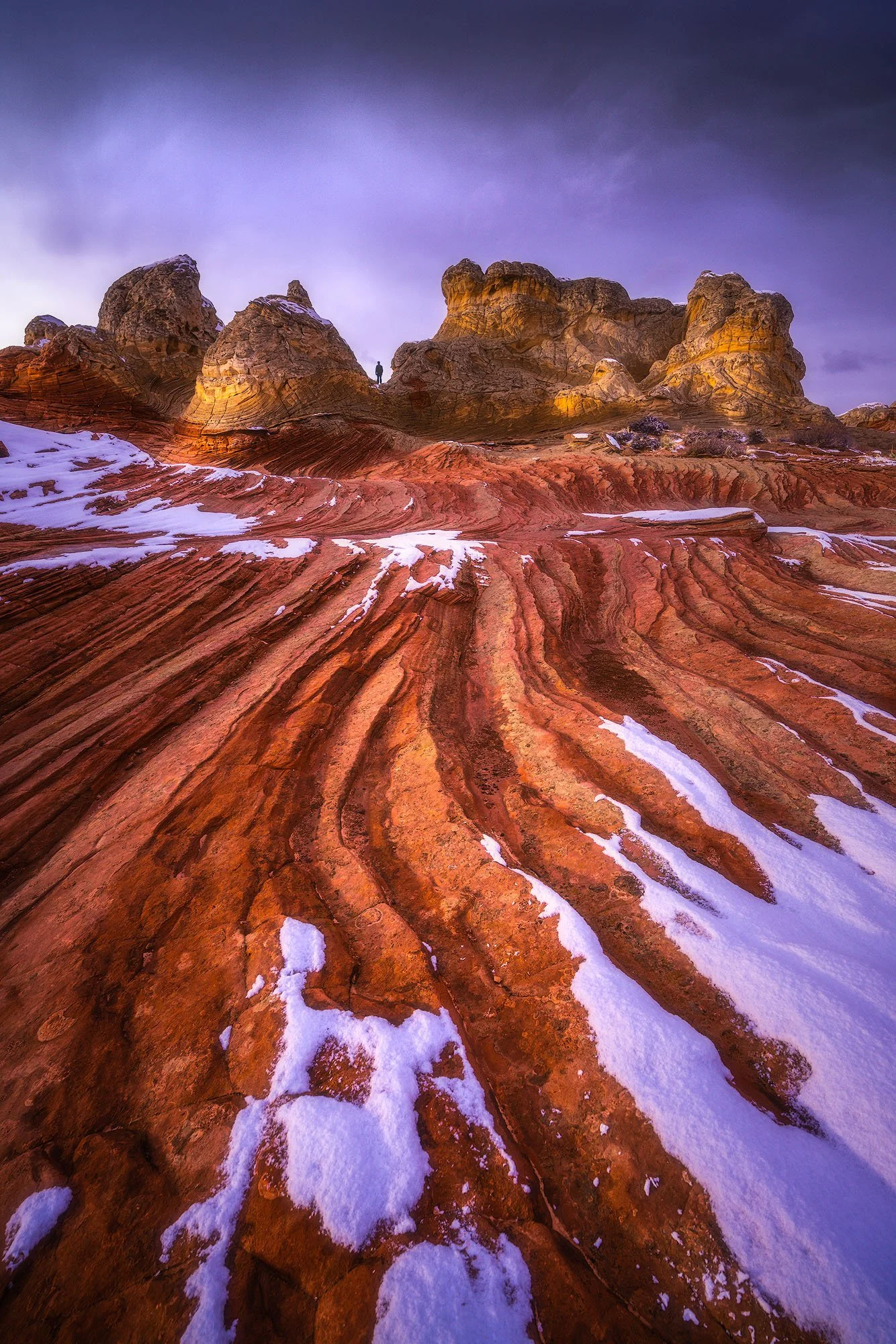 Colorful rock formations with snow patches and a person standing on the rocks under a cloudy sky.