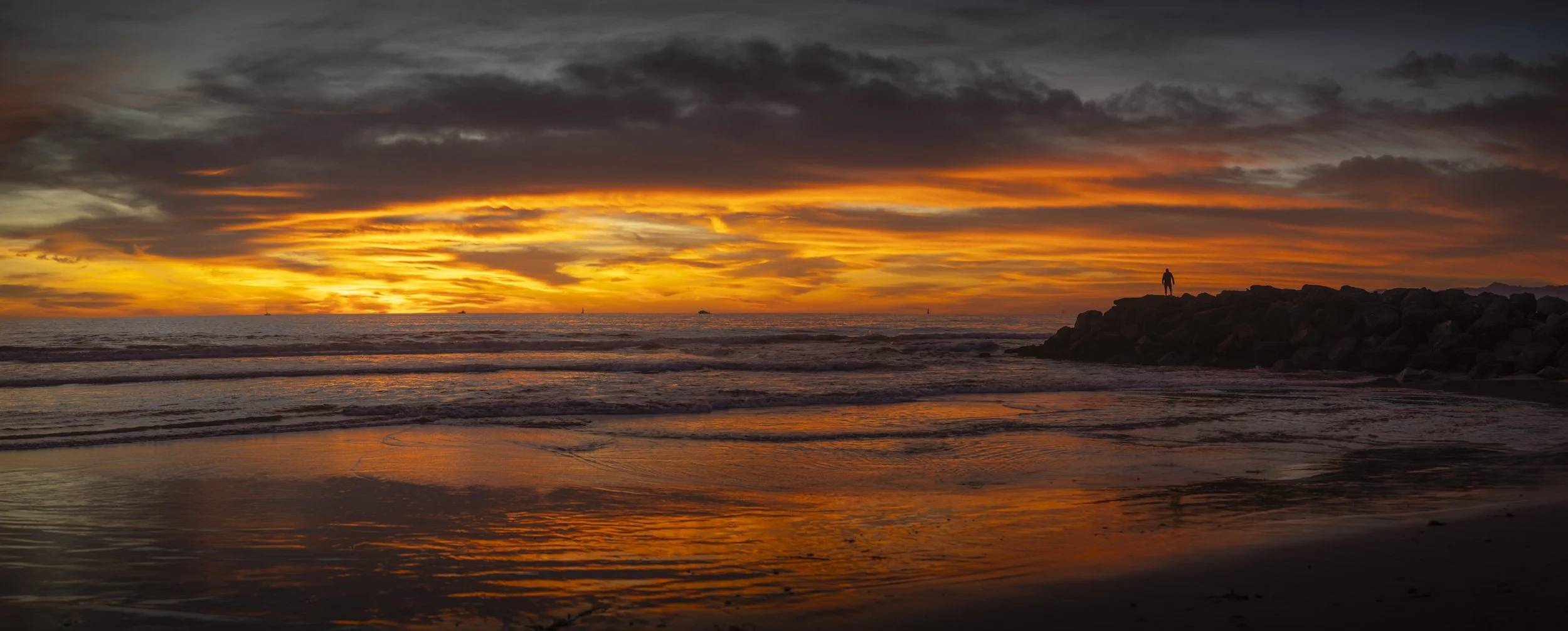 Sunset over the ocean with vibrant orange and yellow hues, dark clouds overhead, and a silhouette of a person standing on a rocky jetty