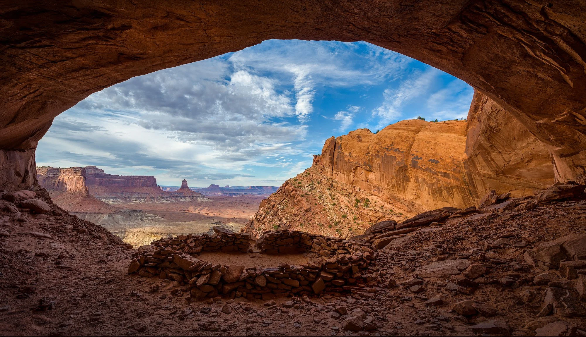 View of a desert canyon landscape with rock formations, viewed from inside a cave or alcove, with a partly cloudy sky overhead.