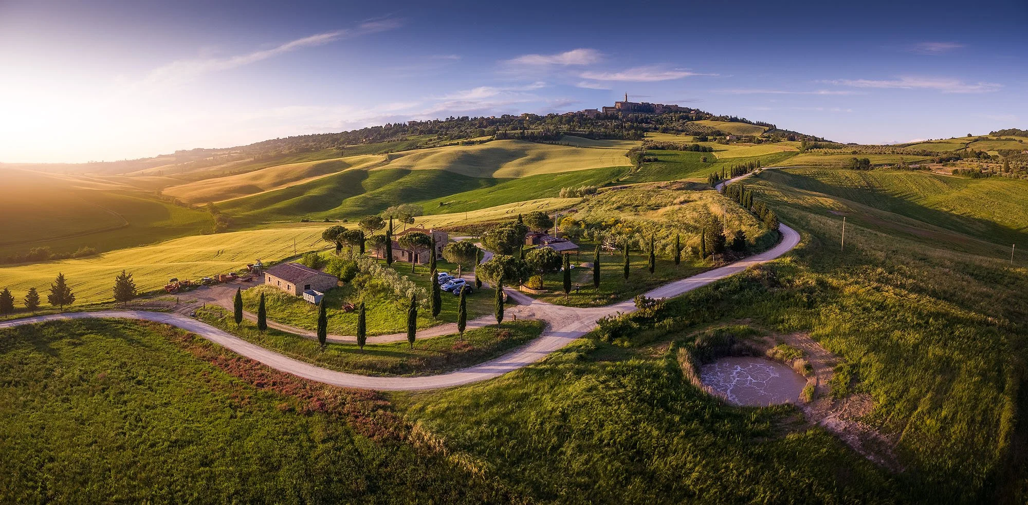 Sunset over rolling green hills with winding roads, cypress trees, and a small pond in a rural landscape.