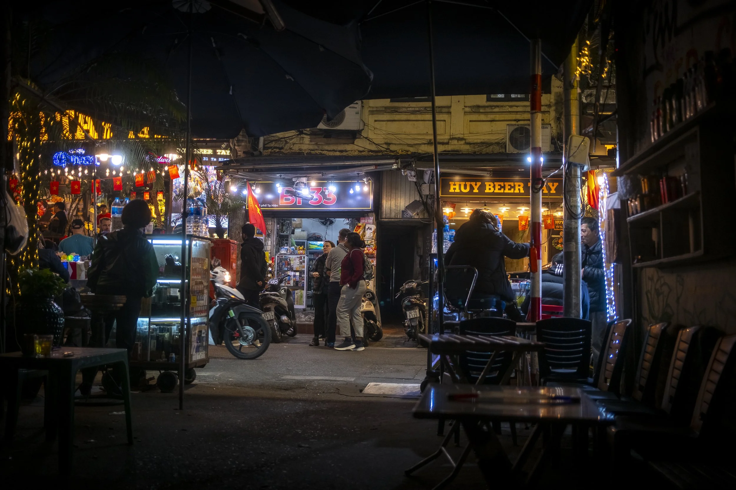 Night view of a busy street with small shops and eateries decorated with string lights, people walking and sitting, motorcycles parked, and neon signs, including a bar called 'B. 33' and a stall named 'Huy Beer'.