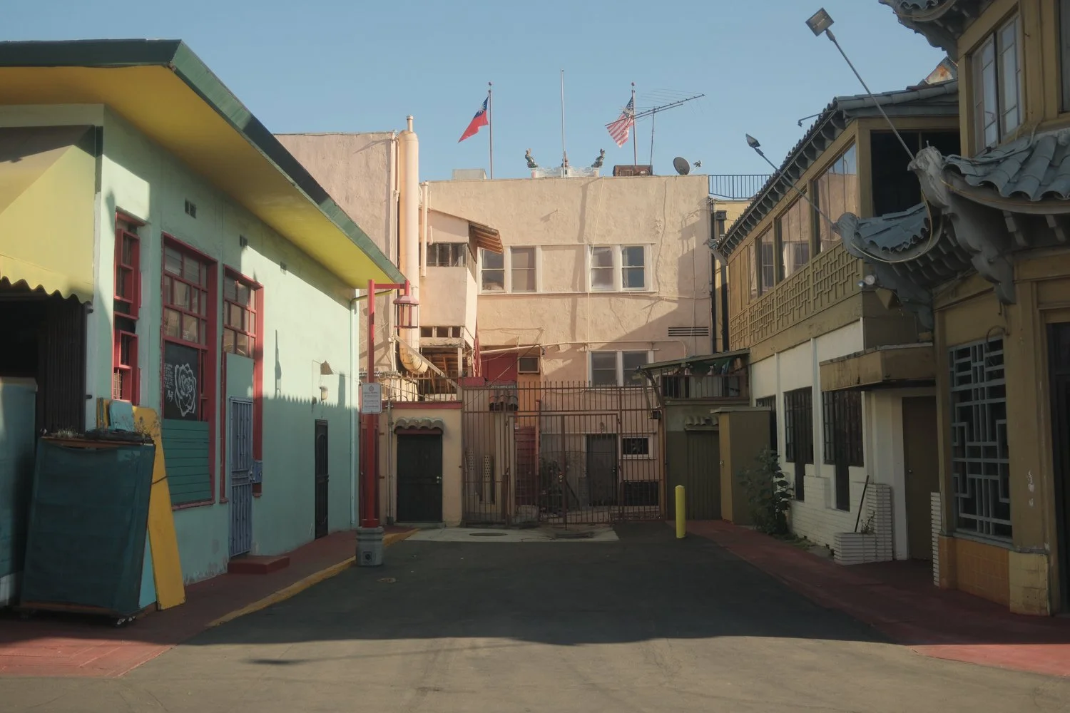 Alleyway with colorful buildings, potted plants, and flags on rooftops, sunny day.