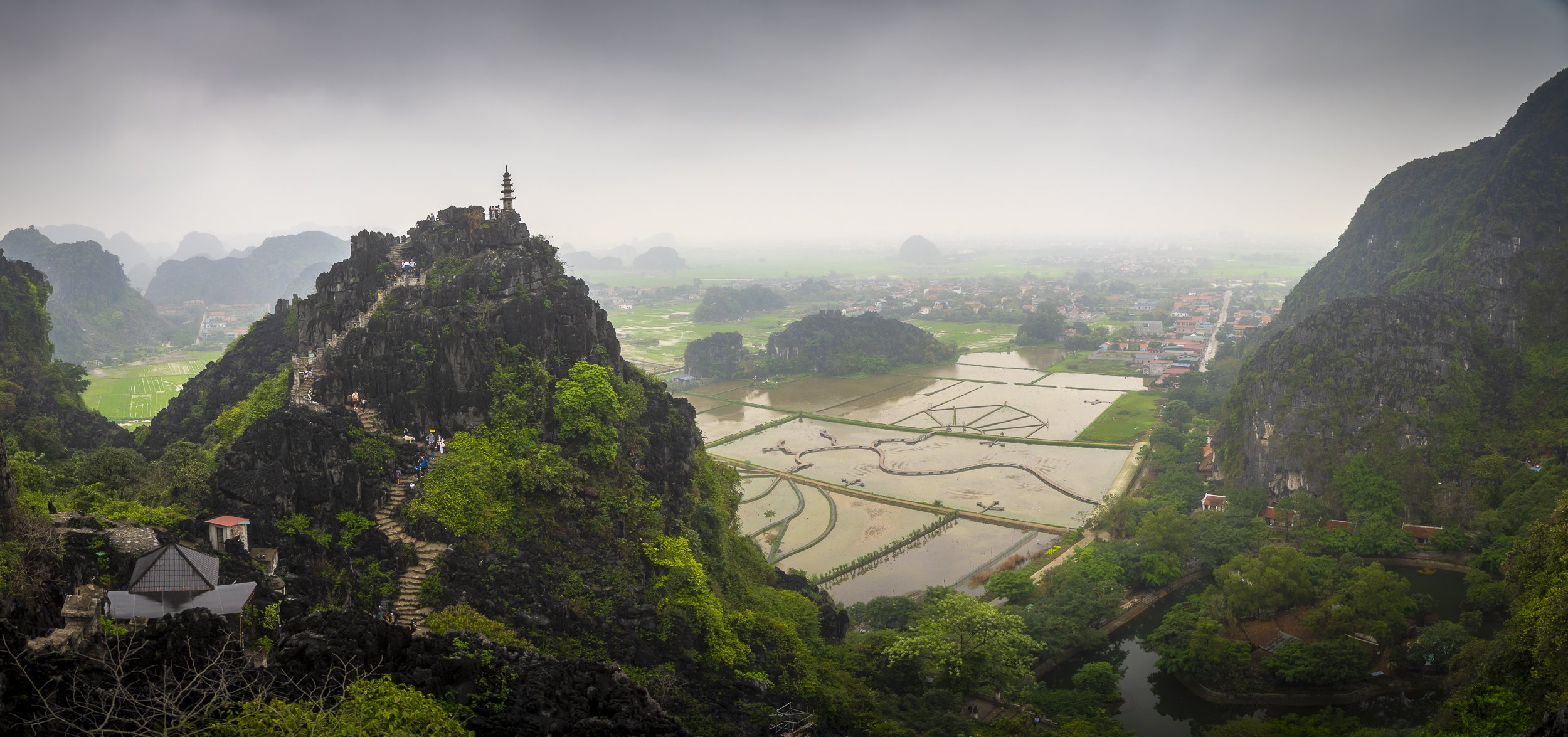 A mountainous landscape with a pagoda on top of the left mountain and terraced rice fields below, surrounded by lush green trees and a small river, with a town in the background under an overcast sky.