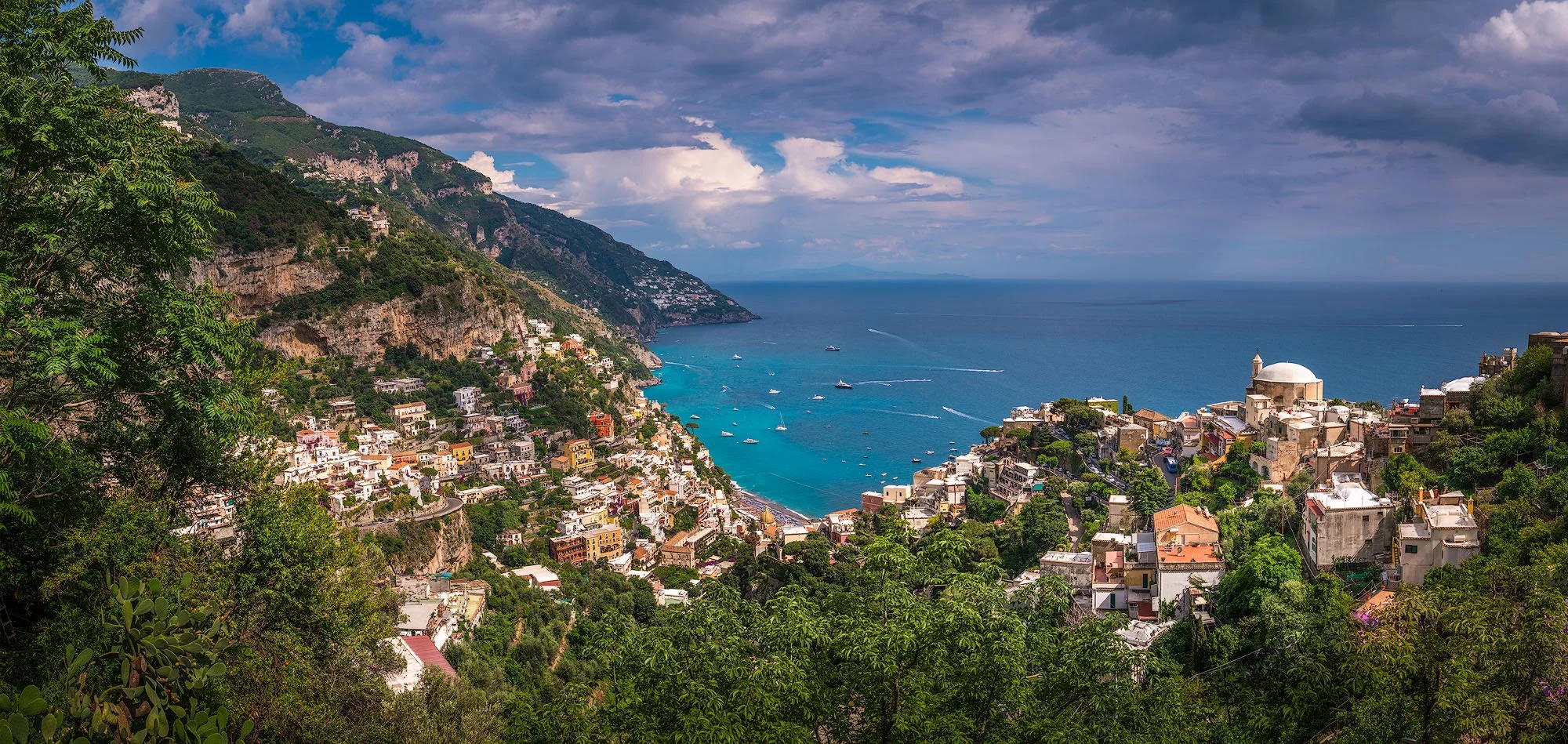 A coastal town built on steep hills with colorful buildings, a large white-domed church, and the blue sea with boats, in a Mediterranean landscape