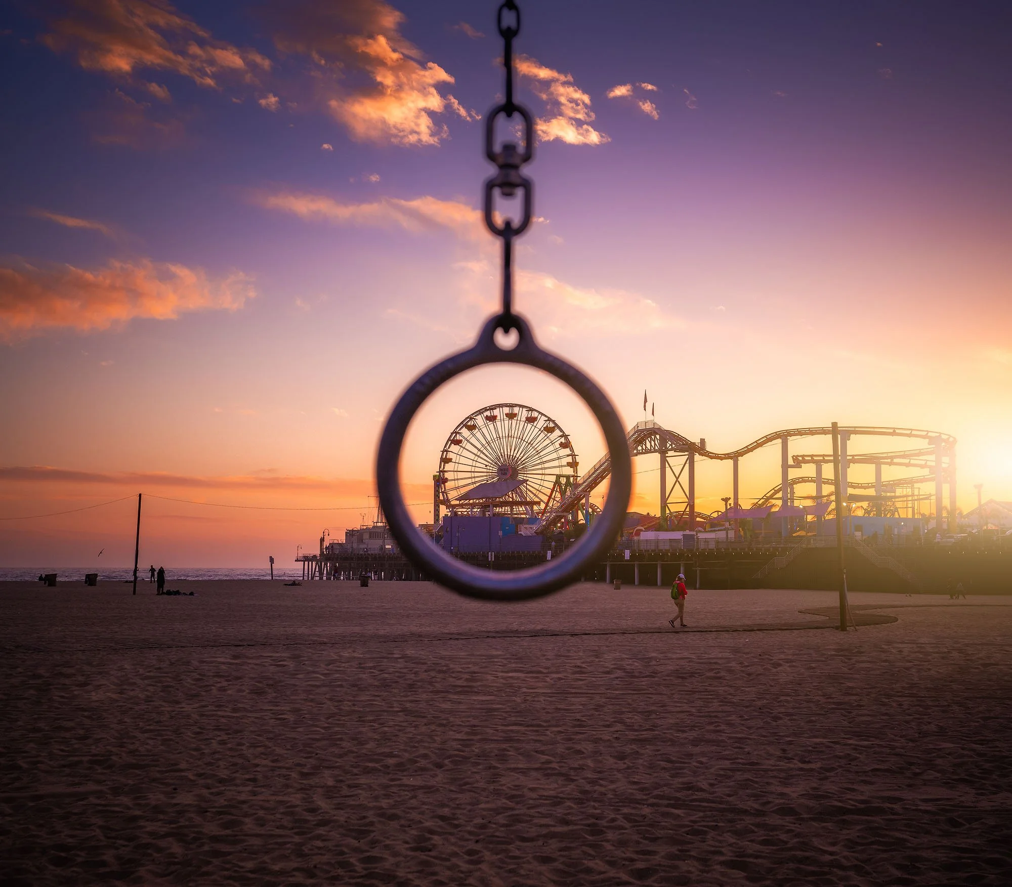A sunset at an amusement park on the beach with a swing in the foreground, a roller coaster and a Ferris wheel in the background.