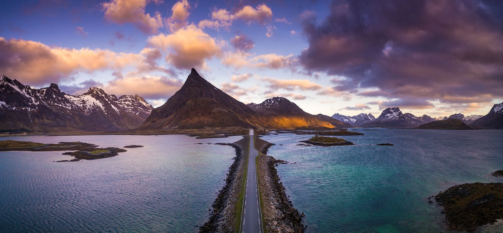 Scenic view of a mountain landscape with a narrow road running through a waterbody, surrounded by snow-capped peaks and a colorful sky.