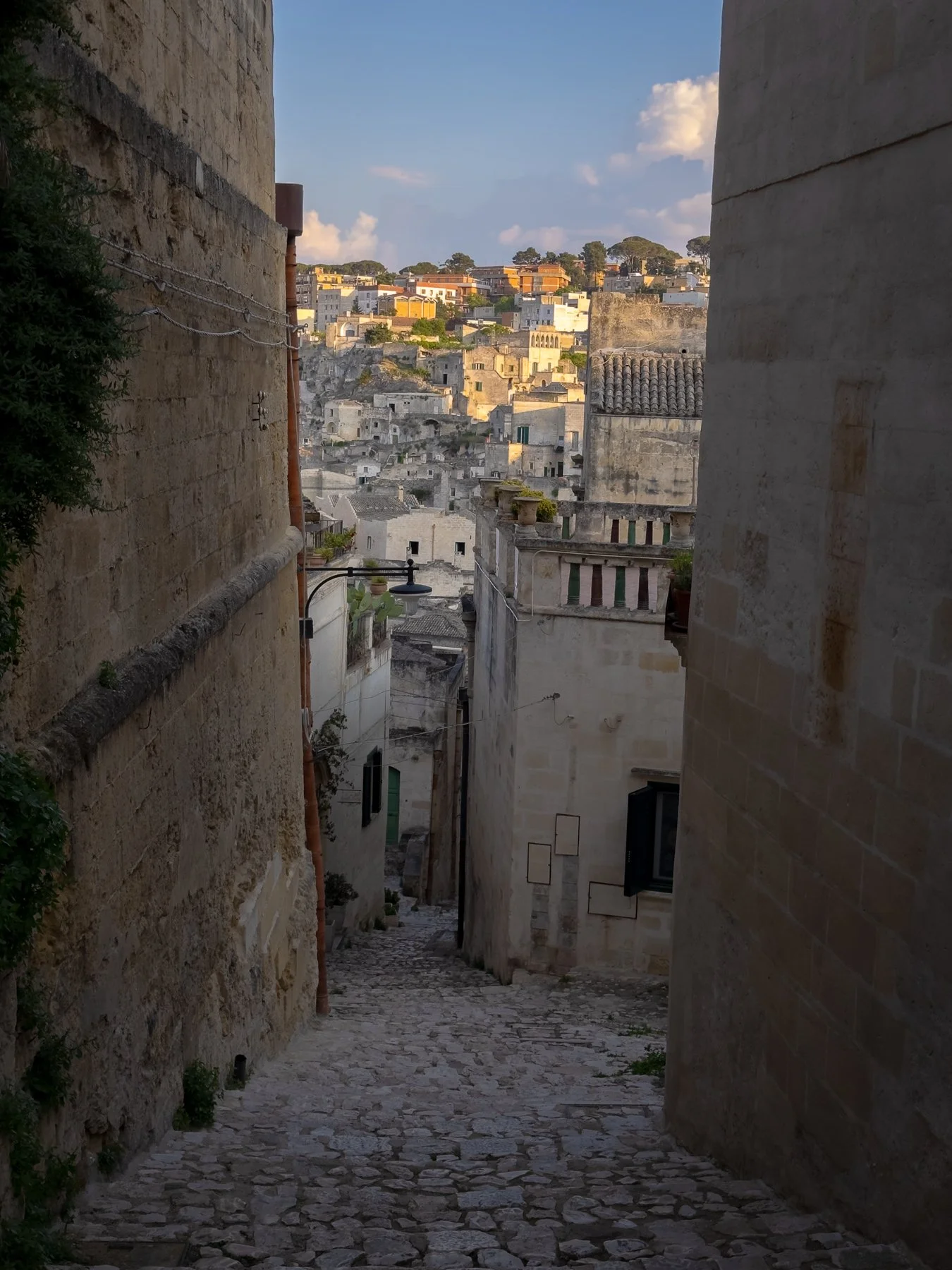 Narrow cobblestone alleyway between old stone buildings leading to a hillside town with colorful buildings under a blue sky.