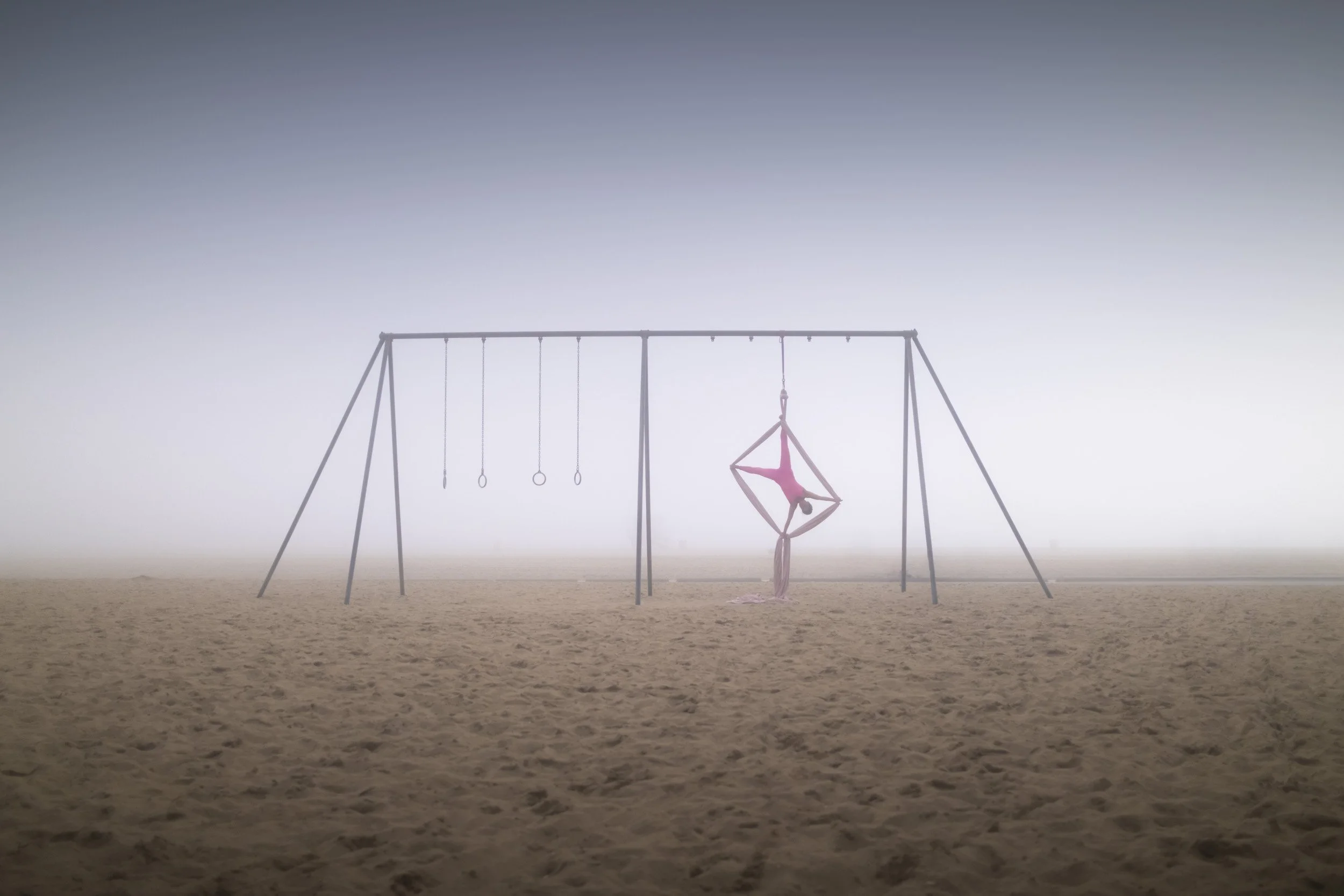 A person practicing aerial silk yoga on a swing set in a foggy beach setting.