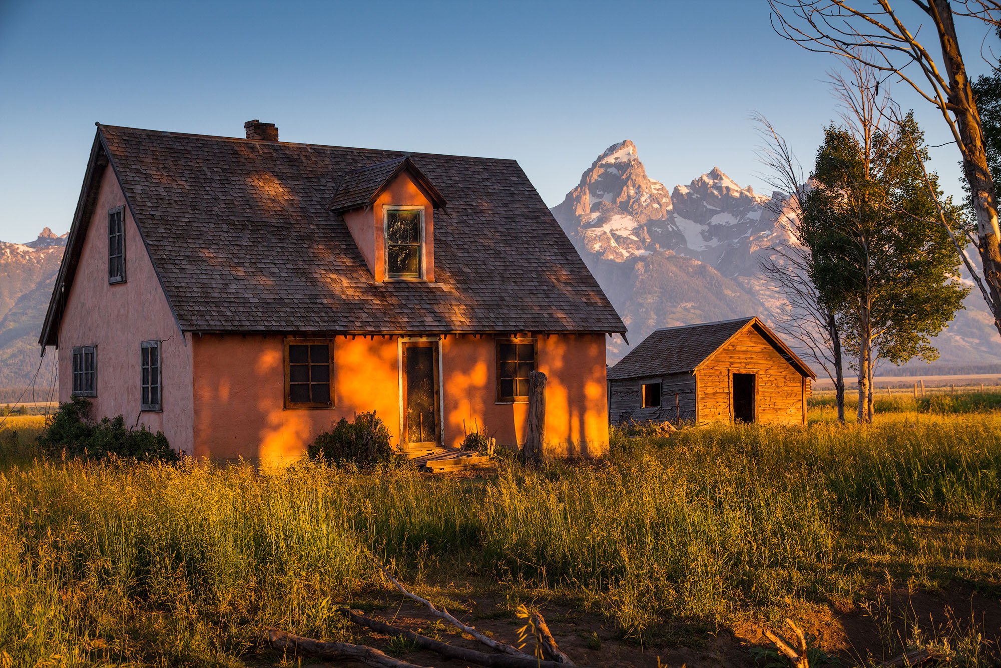 A rustic farmhouse with pinkish walls and a shingle roof, illuminated by warm evening sunlight, with a small wooden shed nearby, surrounded by tall grass and a few trees, and snow-capped mountains in the background.