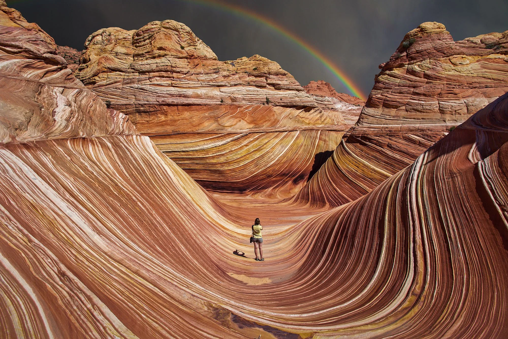 A person standing in a colorful, wavy rock landscape with a rainbow in the dark sky overhead.