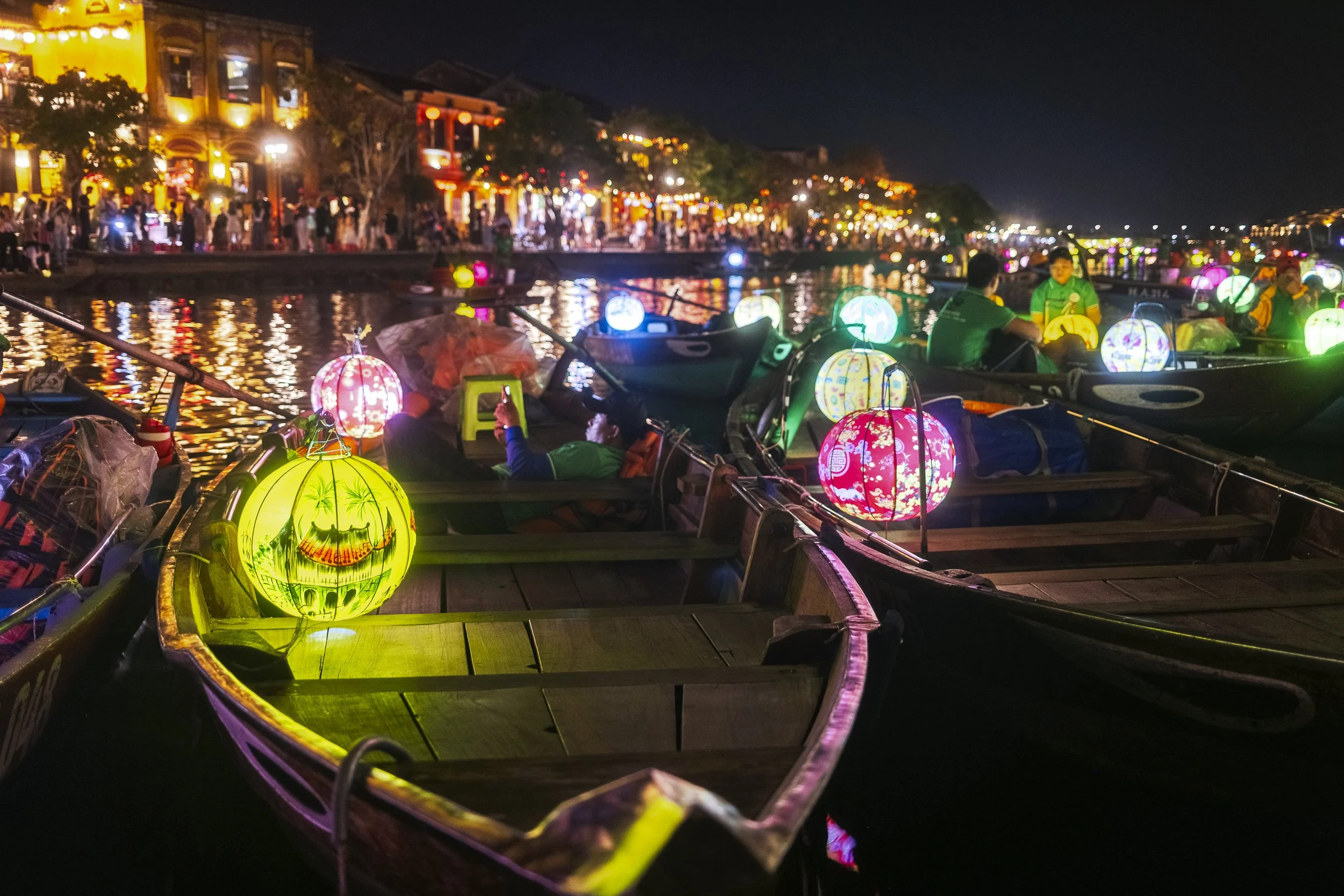 Night scene of colorful lanterns illuminating boats on a river, with a lively riverside street filled with people and lit-up buildings in the background.