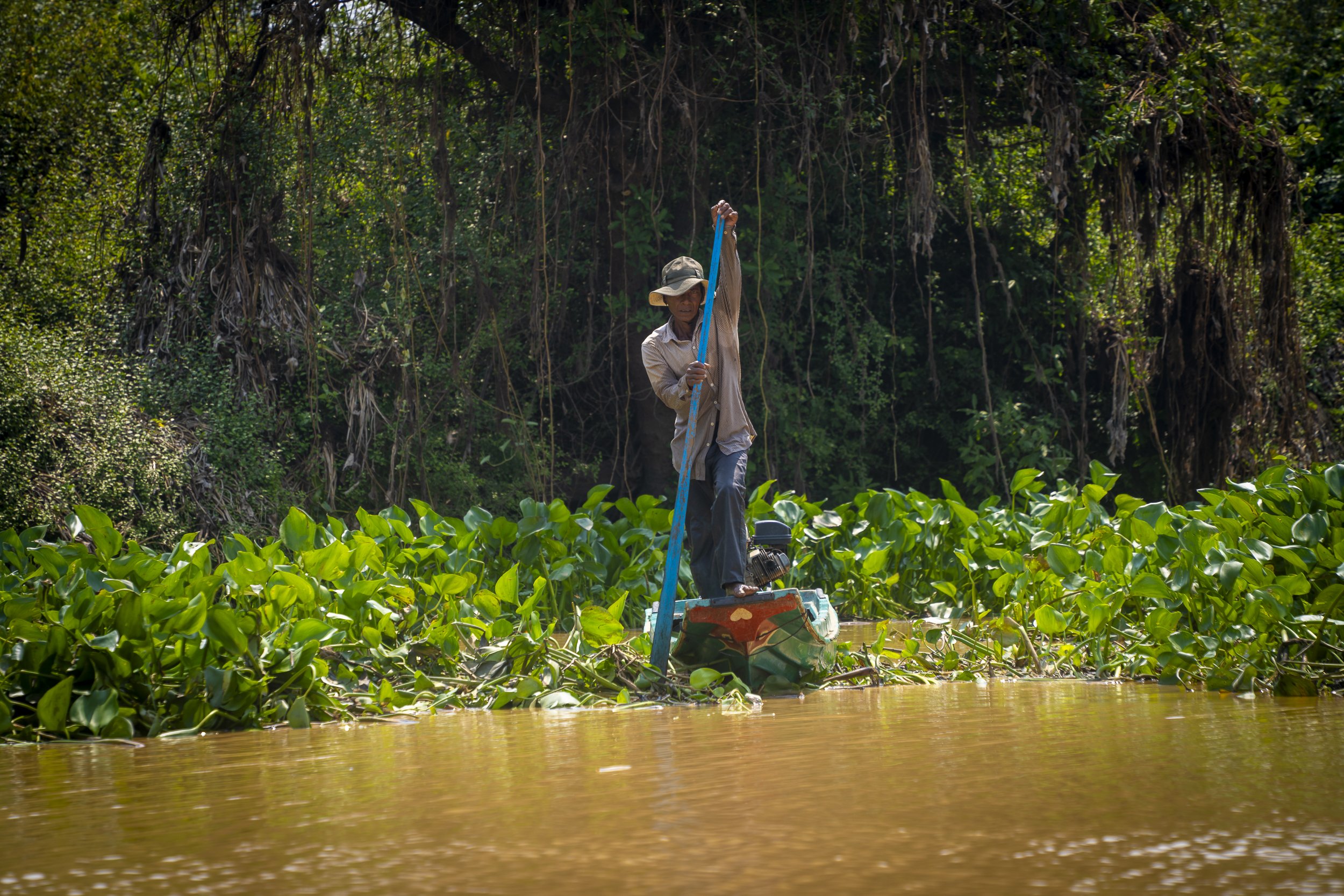 A man standing on a small boat in a waterway, surrounded by lush green vegetation, holding a long pole used for navigation or fishing.
