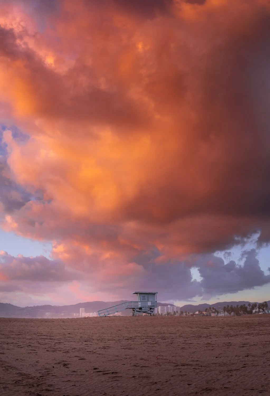 A lifeguard tower on a sandy beach under a dramatic orange and purple sunset sky with scattered clouds.