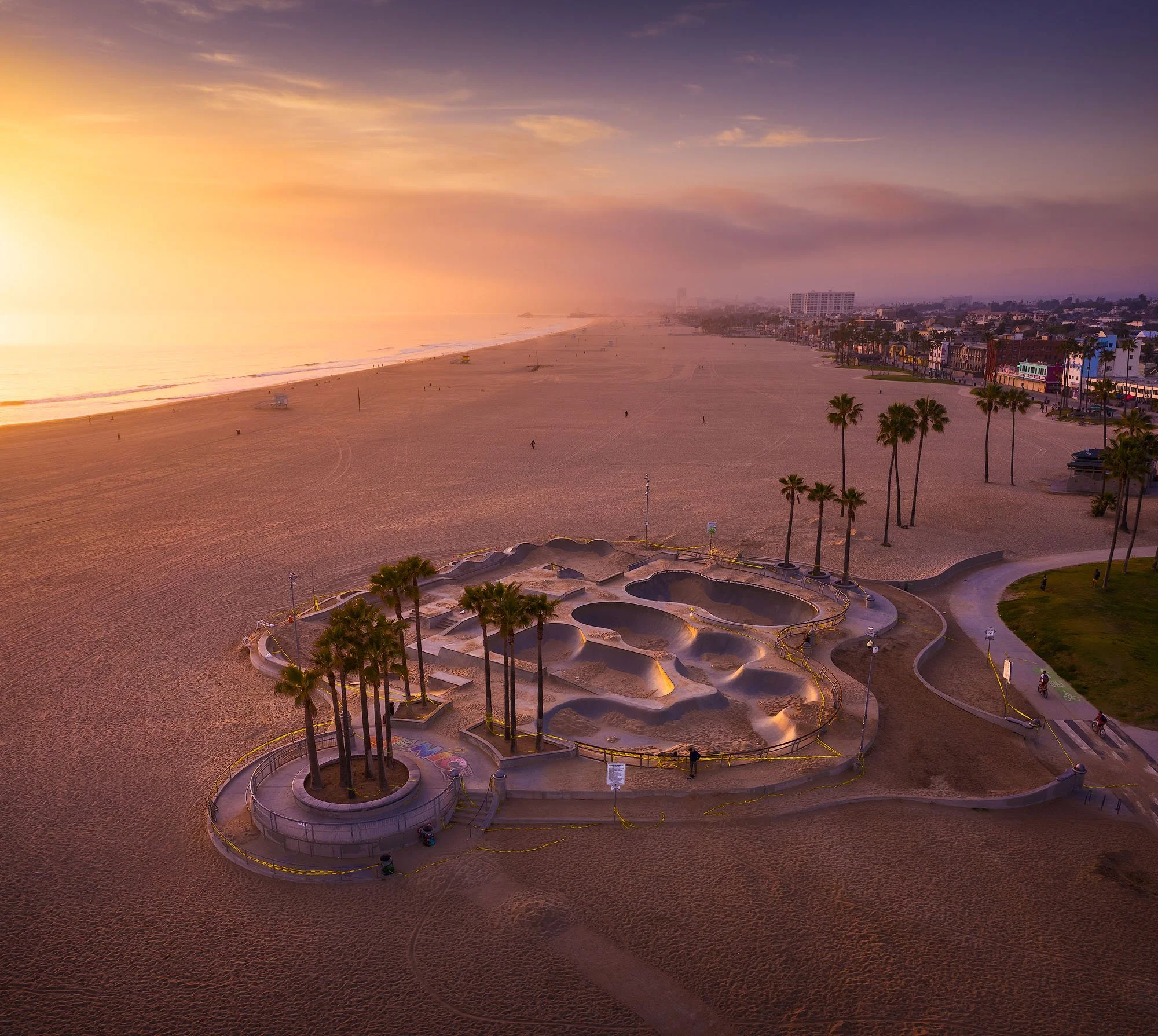 Empty skatepark with multiple ramps and curved concrete features, surrounded by palm trees and set on a sandy beach at sunset with the ocean and distant buildings in the background.